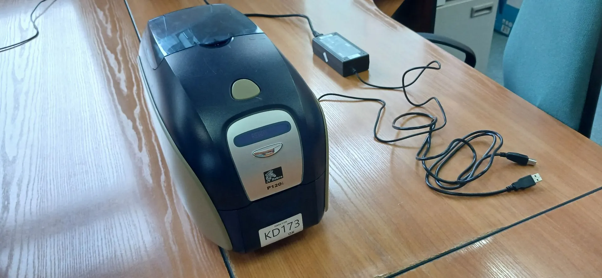 Urządzenie biurowe, prawdopodobnie drukarka etykiet marki Zebra model P120i, w kolorze czarnym i srebrnym, stojące na drewnianym stole. Do urządzenia podłączony jest zasilacz oraz kabel USB. Na obudowie znajduje się biała naklejka z kodem KD173 oraz napisem OK.