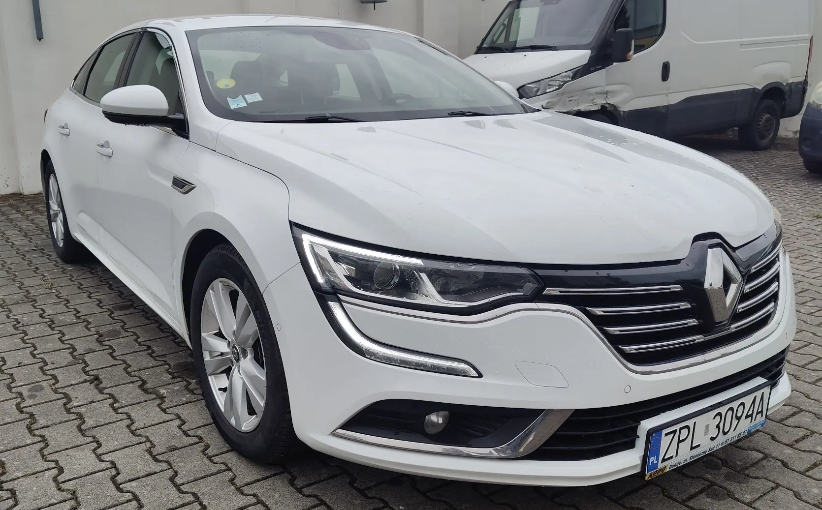 Biały samochód osobowy marki Renault zaparkowany na kostce brukowej. Widoczny jest przód i lewy bok pojazdu. Samochód ma tablicę rejestracyjną o numerze ZPL 3094A. W tle widoczny jest biały furgon.