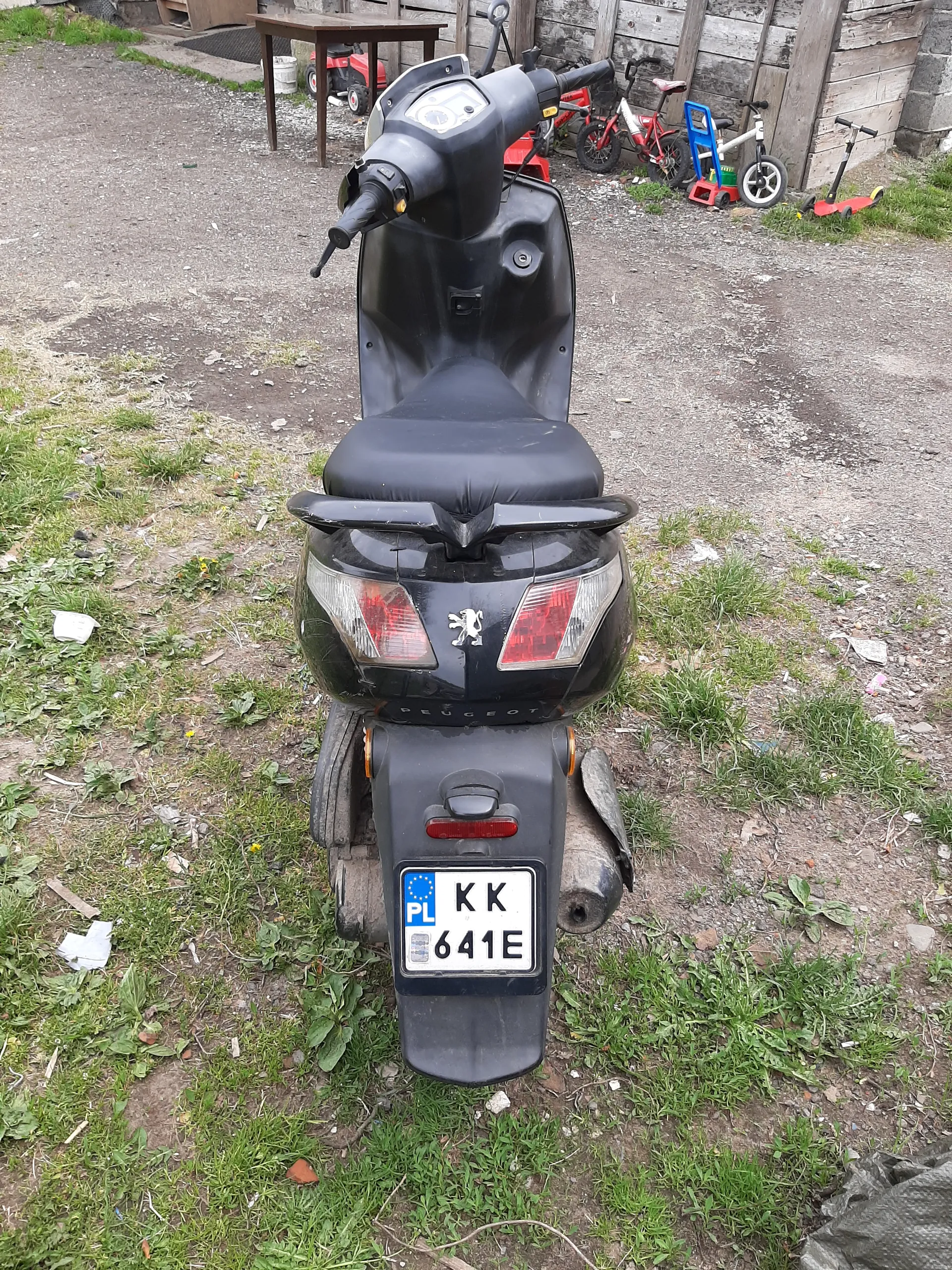 Czarny skuter marki Peugeot z tablicą rejestracyjną KK 641E (PL) zaparkowany na podwórku z trawą i drobnymi śmieci. W tle widoczne są dziecięce rowery (czerwony, niebieski) oraz czerwona hulajnoga. Po lewej stronie drewniana konstrukcja.