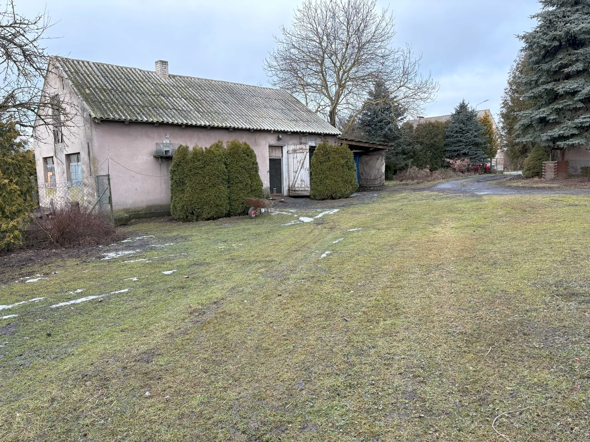 Stara, jednopiętrowa budowla o beżowych ścianach i dachu z blachy falistej, częściowo porośniętej mchem. Przed budynkiem rosną trzy gęste, zielone krzewy, a obok nich stoi rdzewiejący wózek ogrodniczy. Na trawniku z widocznymi plamami śniegu rozciąga się droga prowadząca w głąb posesji. W tle widoczne są drzewa bez liści, iglaste drzewa oraz drugi budynek. Niebo jest zachmurzone. Po lewej stronie budynku znajduje się siatka ogrodzeniowa z bramą.