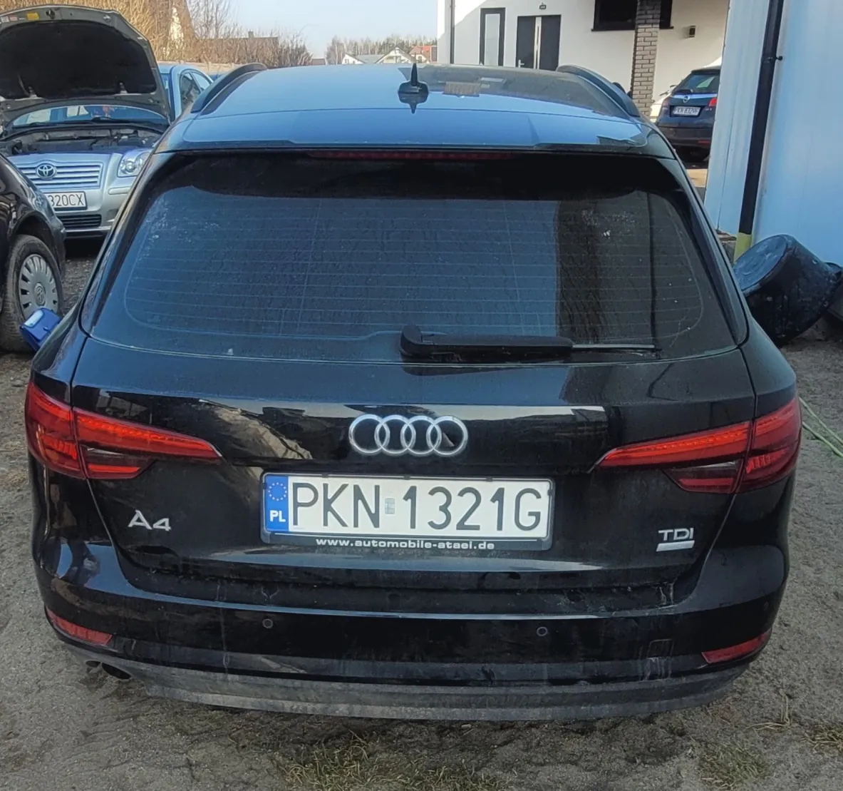Tył czarnego samochodu Audi A4 z tablicą rejestracyjną PL PKN 1321G i adresem www.automobile-atael.de. Widoczne oznaczenia 'A4' po lewej stronie, 'TDI' po prawej oraz logo Audi. W tle inne pojazdy, w tym srebrny samochód z otwartą maską i budynki.