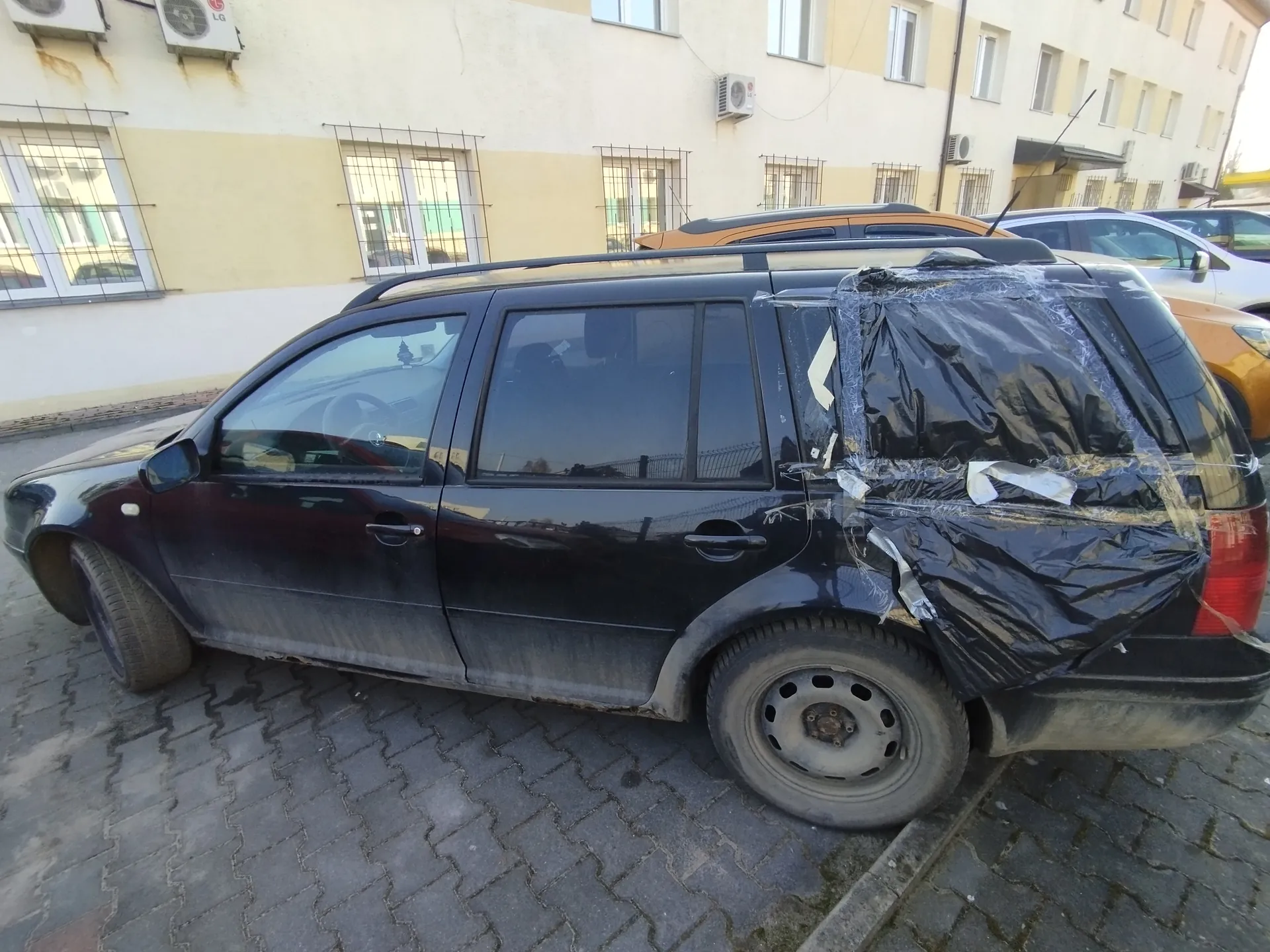 Czarny samochód typu kombi zaparkowany na brukowanej powierzchni. Tylna część pojazdu (w tym szyba) jest przykryta czarną folią przylepioną taśmą. Samochód jest zabrudzony, szczególnie dolne części karoserii. W tle widoczne są budynki mieszkalne z żółto-białą elewacją, oknami z metalowymi kratami oraz klimatyzatorami marki LG. Za czarnym samochodem stoją inne pojazdy: pomarańczowy i srebrny. Na budynkach widoczne są również zewnętrzne jednostki klimatyzacyjne z napisem LG.