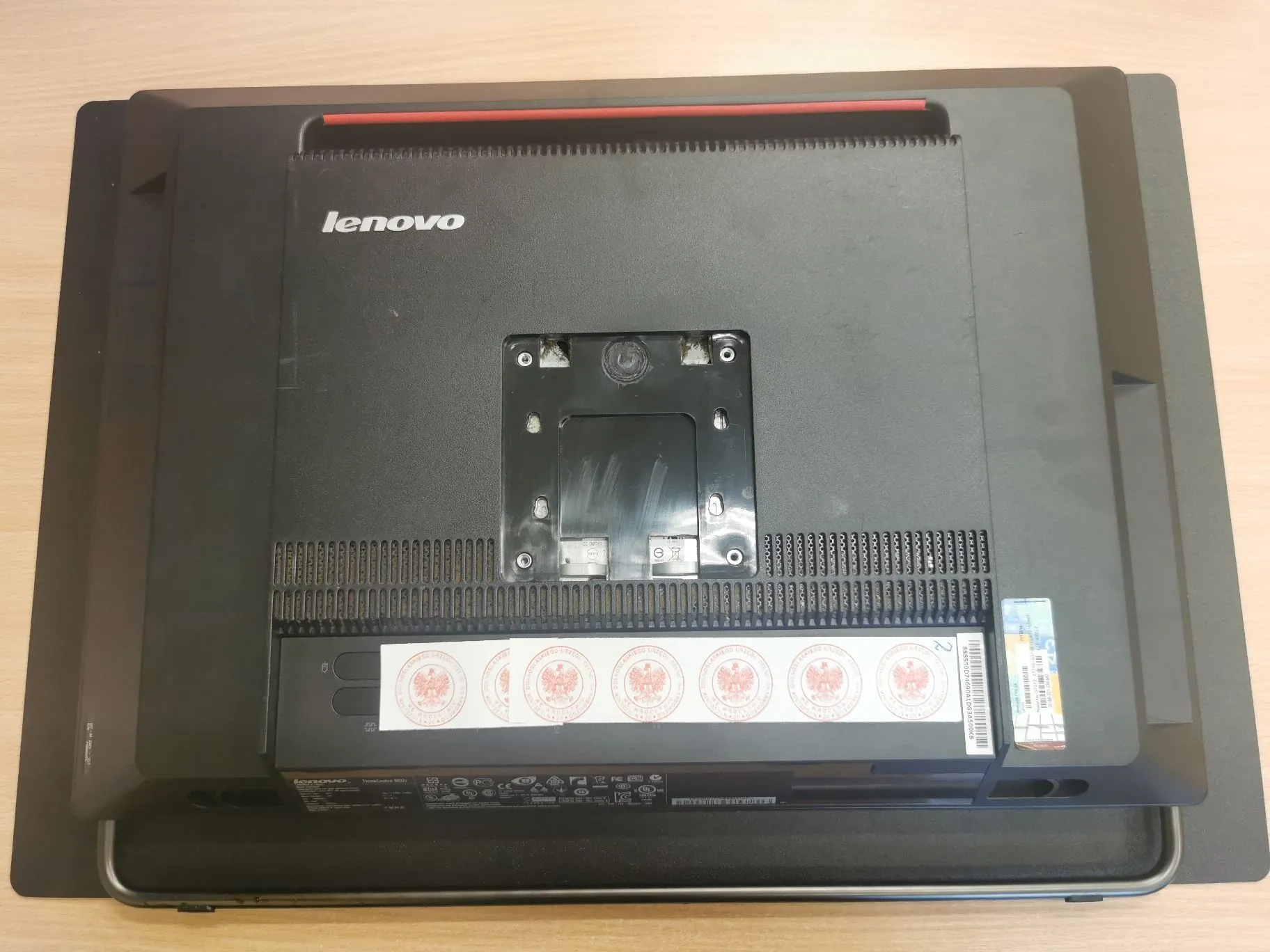 Tylna obudowa monitora marki Lenovo w kolorze czarnym. Na górze znajduje się biały napis 'lenovo'. W centralnej części widoczny jest metalowy uchwyt VESA do montażu na ścianie lub ramieniu. W dolnej części obudowy znajduje się poziomy pasek z kilkoma okrągłymi, czerwono-białymi naklejkami z godłem Polski oraz tabliczka znamionowa z danymi technicznymi i kodami kreskowymi.