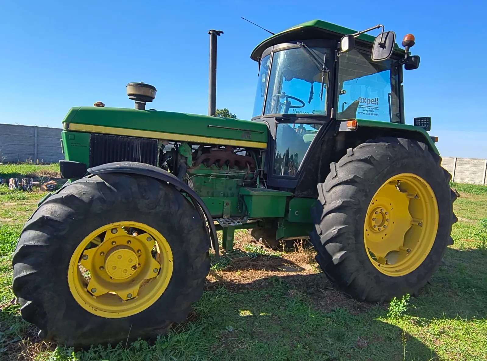 Zielony traktor z żółtymi felgami, zaparkowany na trawiastym terenie na tle niebieskiego nieba. Pojazd posiada zamkniętą kabinę z szybami, w których odbija się otoczenie. Na tylnej szybie widoczna jest naklejka z napisem 'expell'.
