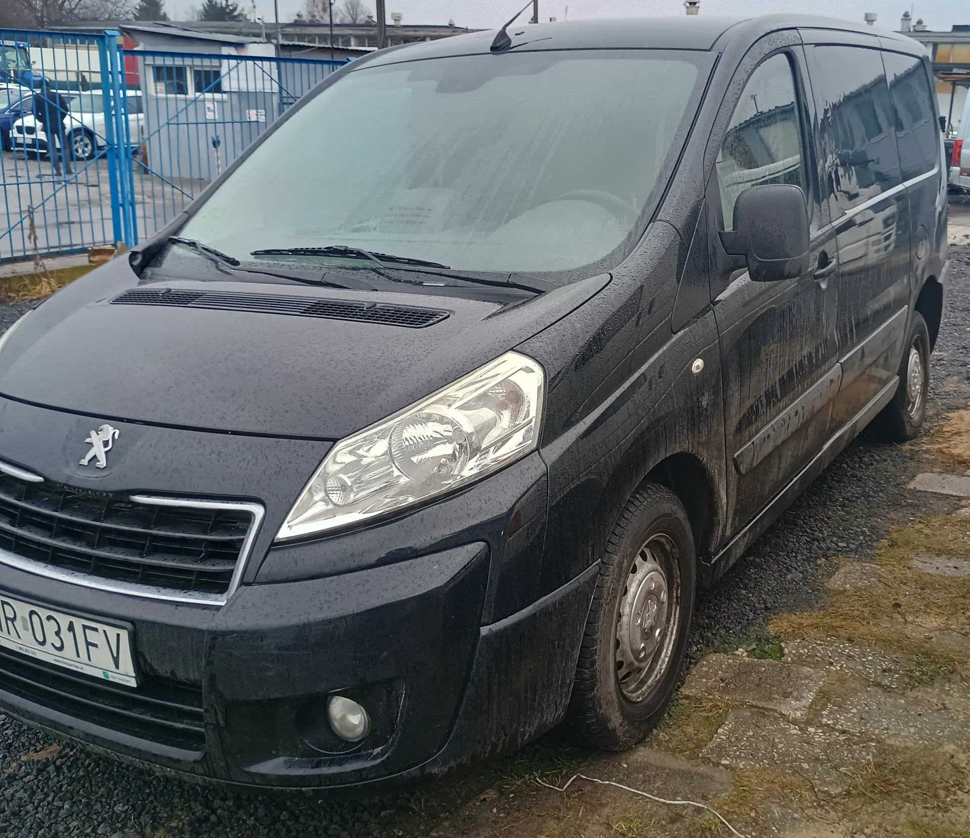 Czarny samochód dostawczy marki Peugeot z widocznym logo na masce, stojący na podwórku z żwirem i betonowymi płytami. Na przodzie tablica rejestracyjna z białym tłem i czarnym napisem R 031FV. Samochód jest zabrudzony, z widocznymi plamami błota i kroplami wody. W tle niebieskie ogrodzenie metalowe, budynki oraz inne pojazdy. Niebo jest zachmurzone.