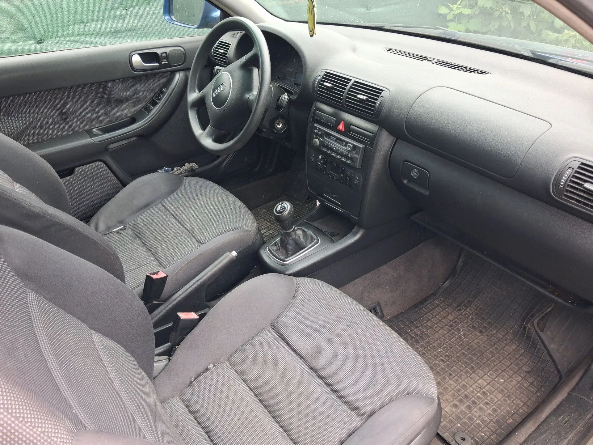 Wnętrze samochodu marki Audi widziane z perspektywy pasażera. Widoczna jest szara tapicerka materiałowa na przednich fotelach, czarna deska rozdzielcza, kierownica z logo Audi, konsola środkowa z radiem i lewarem zmiany biegów oraz gumowe dywaniki na podłodze.