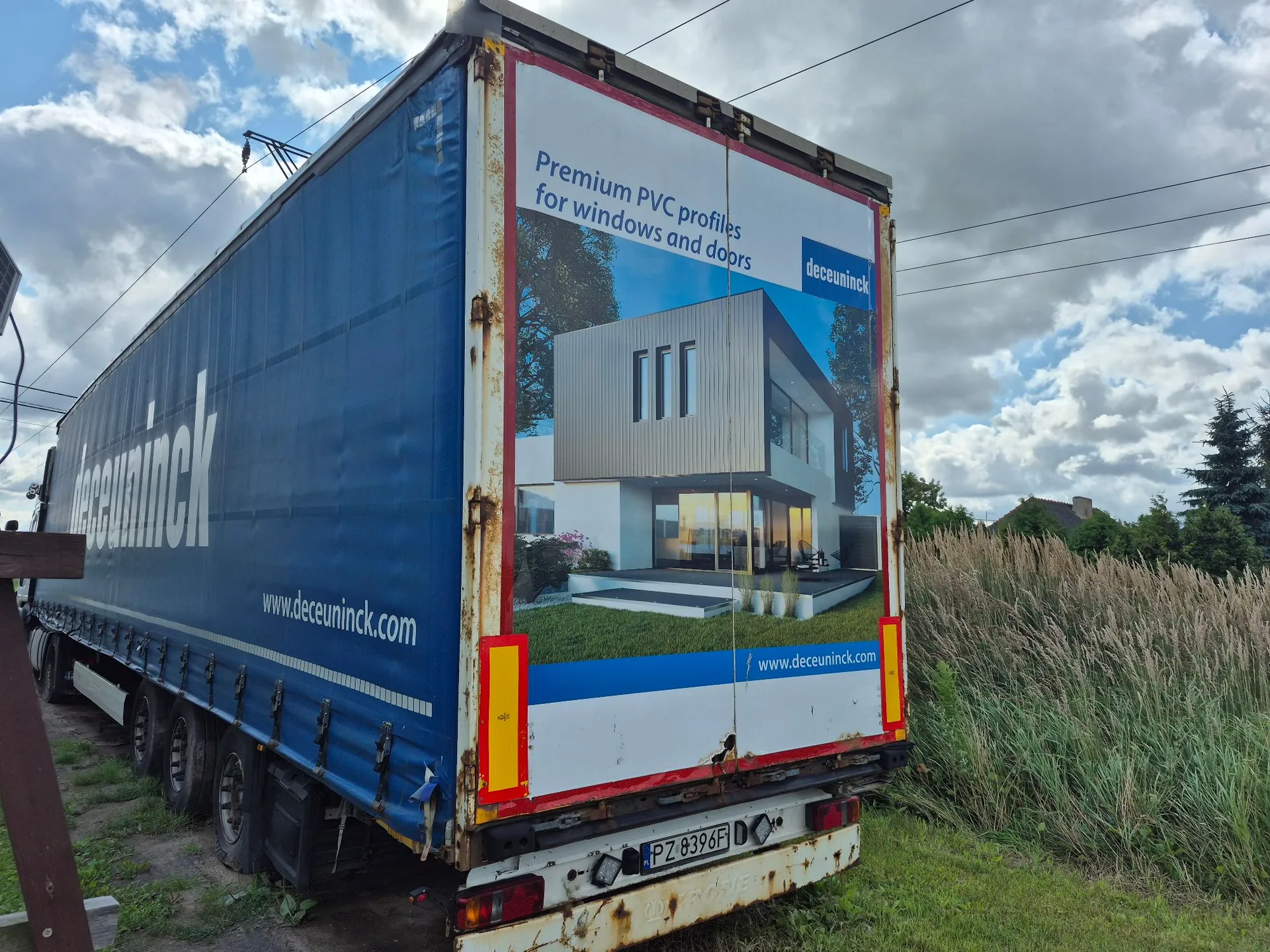 Tylna i boczna część niebieskiej naczepy ciężarówki zaparkowanej na trawie. Na tylnej ścianie naczepy znajduje się reklama z napisem „Premium PVC profiles for windows and doors” oraz logo „deceuninck”. Reklama przedstawia nowoczesny, biały dom z dużymi przeszkleniami. Na dole reklamy widnieje adres strony internetowej „www.deceuninck.com”. Na boku naczepy znajduje się duży biały napis „deceuninck” oraz adres strony internetowej. Pojazd ma polską tablicę rejestracyjną o numerze PZ 8396F.