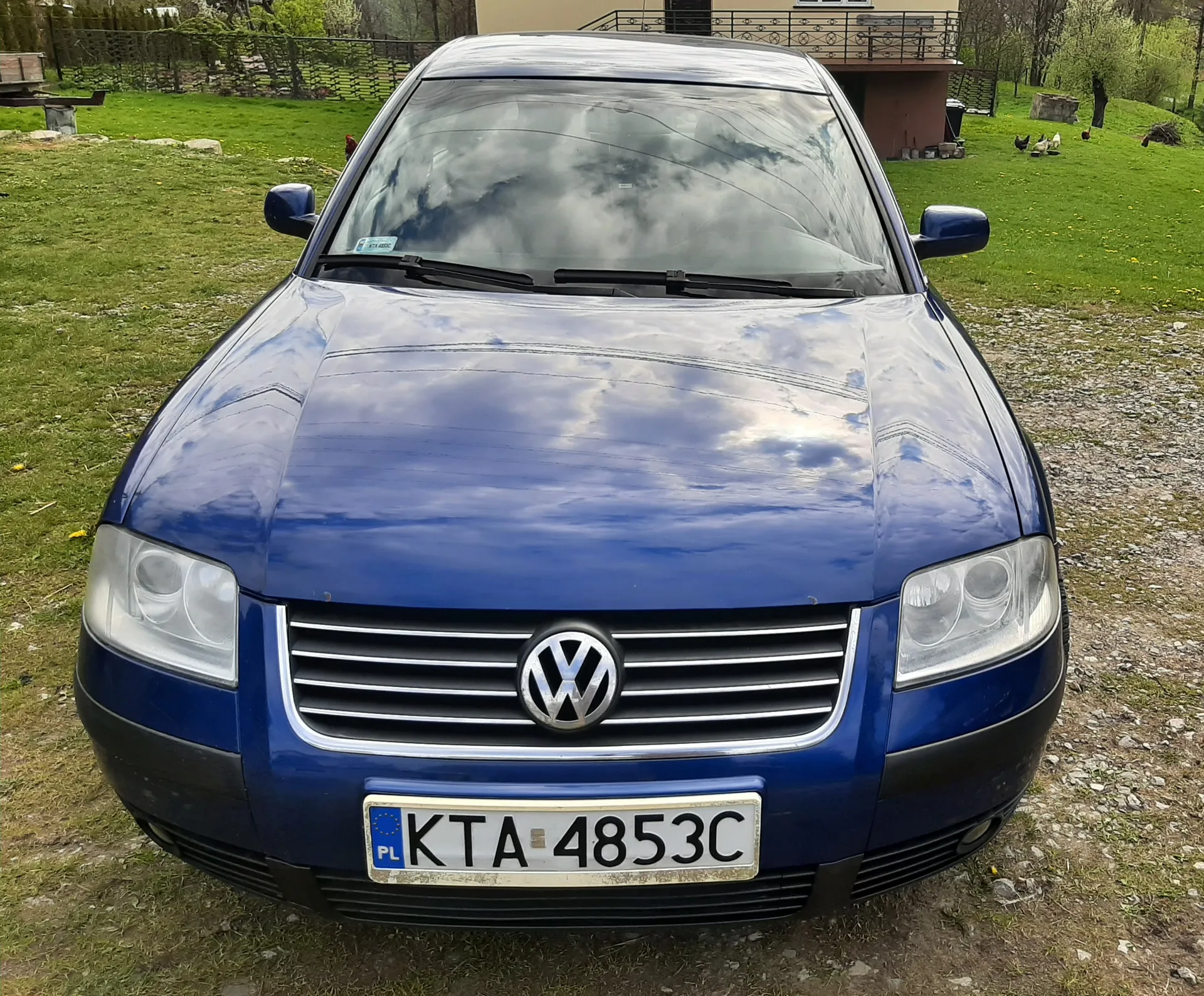 Niebieski samochód marki Volkswagen z przodu, z widocznym logo na atrapie chłodnicy i tablicą rejestracyjną PL KTA 4853C. Samochód stoi na żwirowej powierzchni z zieloną trawą w tle. W tle widoczny jest dom z balkonem, ogrodzenie, drzewa oraz kilka kur na trawie. Na masce odbijają się chmury i niebo.