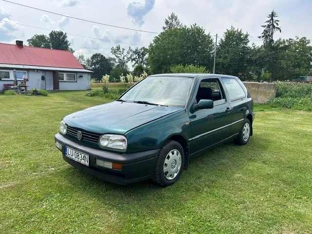 Ciemnozielony samochód Volkswagen Golf trzeciej generacji zaparkowany na zielonej trawie. Auto ma polską tablicę rejestracyjną o numerze LU 0834N. W tle widoczny jest dom z czerwonym dachem oraz drzewa pod jasnym, lekko zachmurzonym niebem.