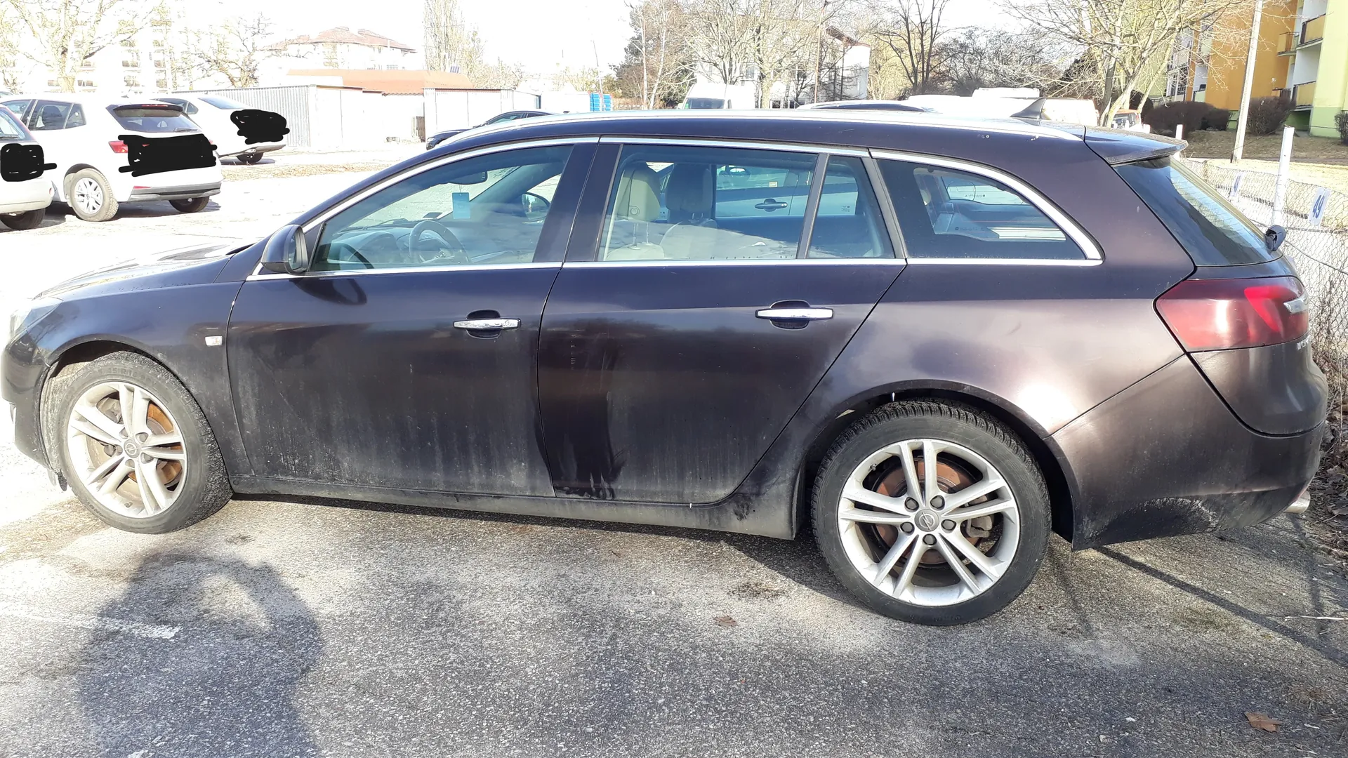 Ciemnoszary samochód typu kombi zaparkowany na asfaltowej powierzchni. Pojazd jest zabrudzony, widoczne są ślady kurzu i brudu na karoserii. W tle znajdują się inne samochody, w tym biały SUV z zamalowanymi fragmentami, budynki mieszkalne, drzewa bez liści oraz ogrodzenie. Na pierwszym planie widoczne są srebrne felgi i hamulce tarczowe. Scena rozgrywa się na otwartym parkingu w słoneczny dzień.