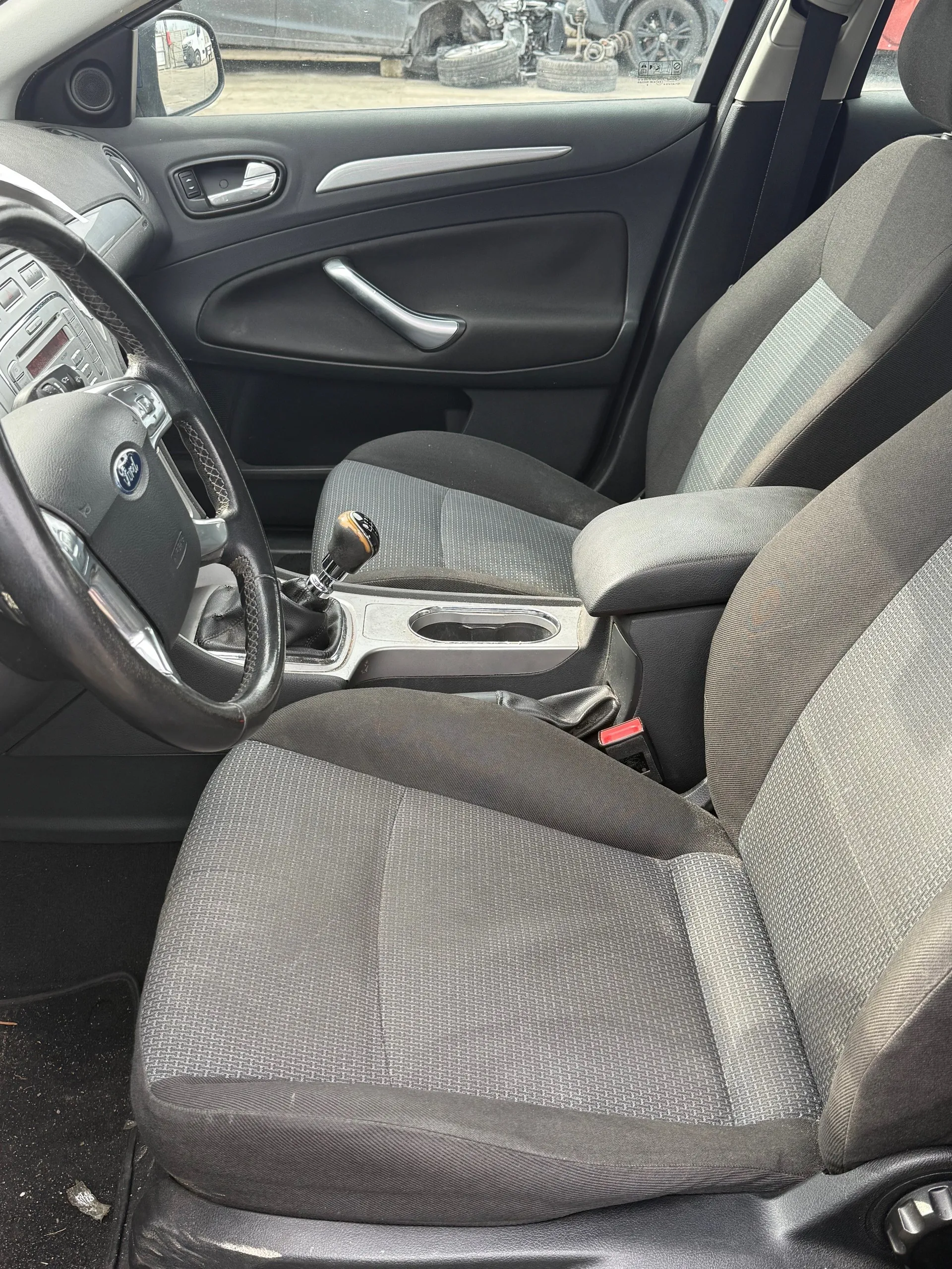 Wnętrze samochodu marki Ford z widocznym kierownicą z logo Ford, szarymi siedzeniami w tkaninie, dźwignią zmiany biegów i panelami drzwiowymi. W tle przez okno widać opony i fragment innego pojazdu na zewnątrz.