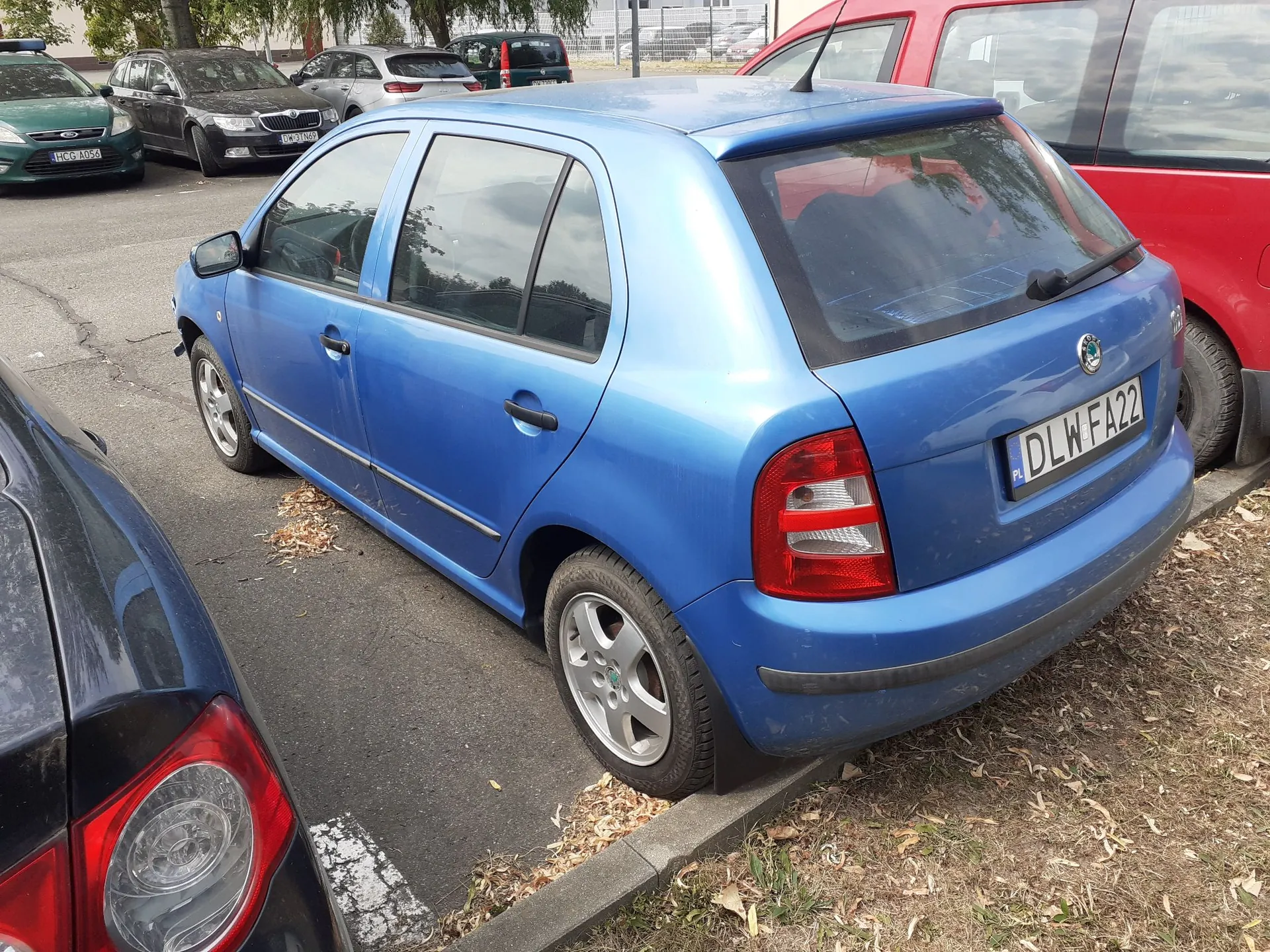 Niebieski samochód marki Volkswagen Polo zaparkowany na parkingu. Widoczny jest tył i lewy bok pojazdu z tablicą rejestracyjną o numerze DLW FA22. W tle znajdują się inne zaparkowane samochody w kolorach zielonym, czarnym, białym i czerwonym.