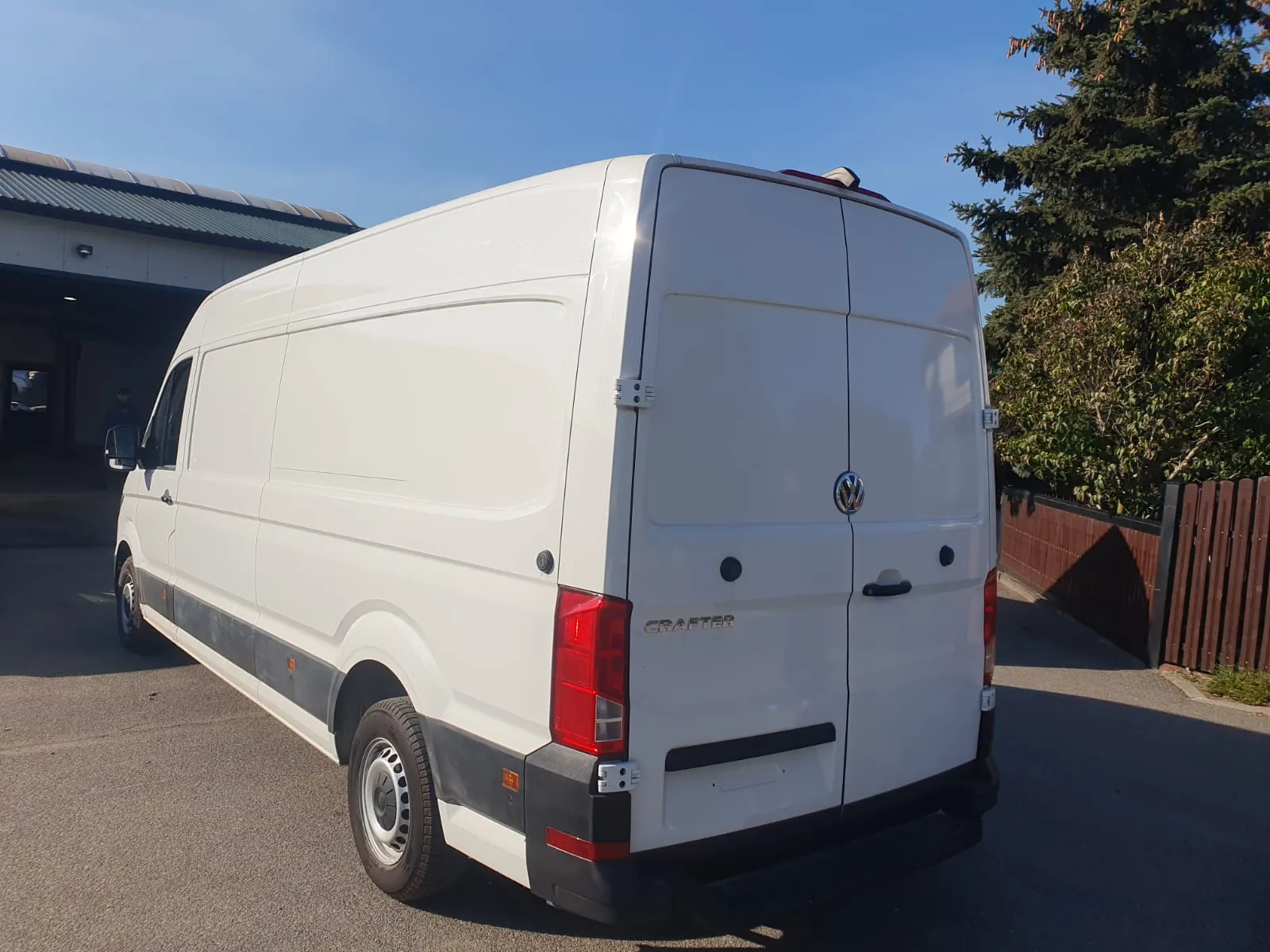 Biały furgon marki Volkswagen Crafter zaparkowany na asfaltowym podjeździe. Pojazd jest widoczny z tyłu i z boku. Na tylnych drzwiach znajduje się logo Volkswagena oraz napis 'CRAFTER'. Dolna część nadwozia jest wykończona szarymi, plastikowymi osłonami. W tle widoczny jest fragment budynku z dachem oraz zielone drzewa pod bezchmurnym, niebieskim niebem.
