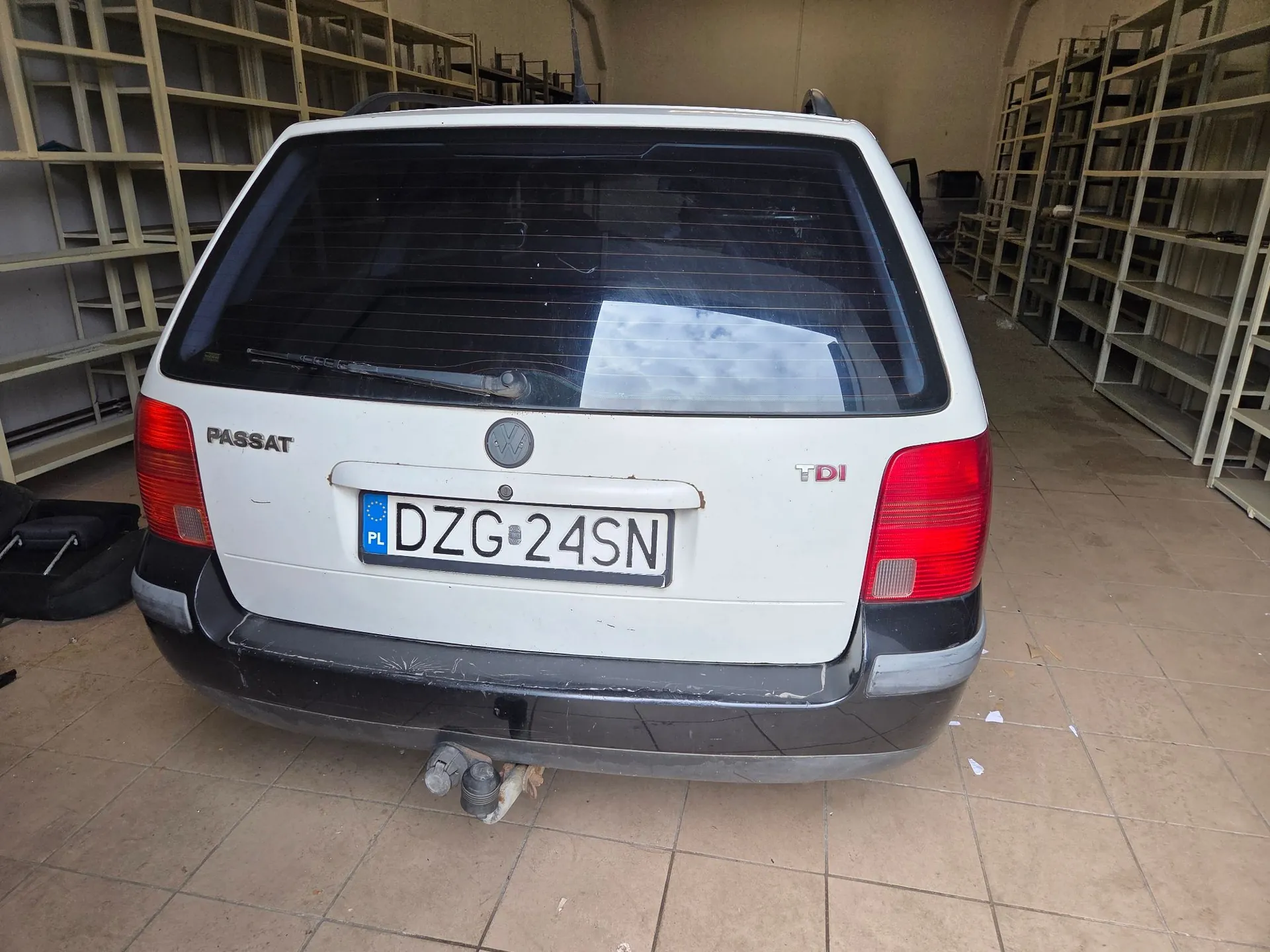 Tylny widok białego samochodu marki Volkswagen Passat TDI z polską tablicą rejestracyjną o numerze DZG 24SN. Samochód znajduje się w pomieszczeniu przypominającym magazyn lub garaż z regałami w tle. Pojazd posiada zamontowany hak holowniczy.