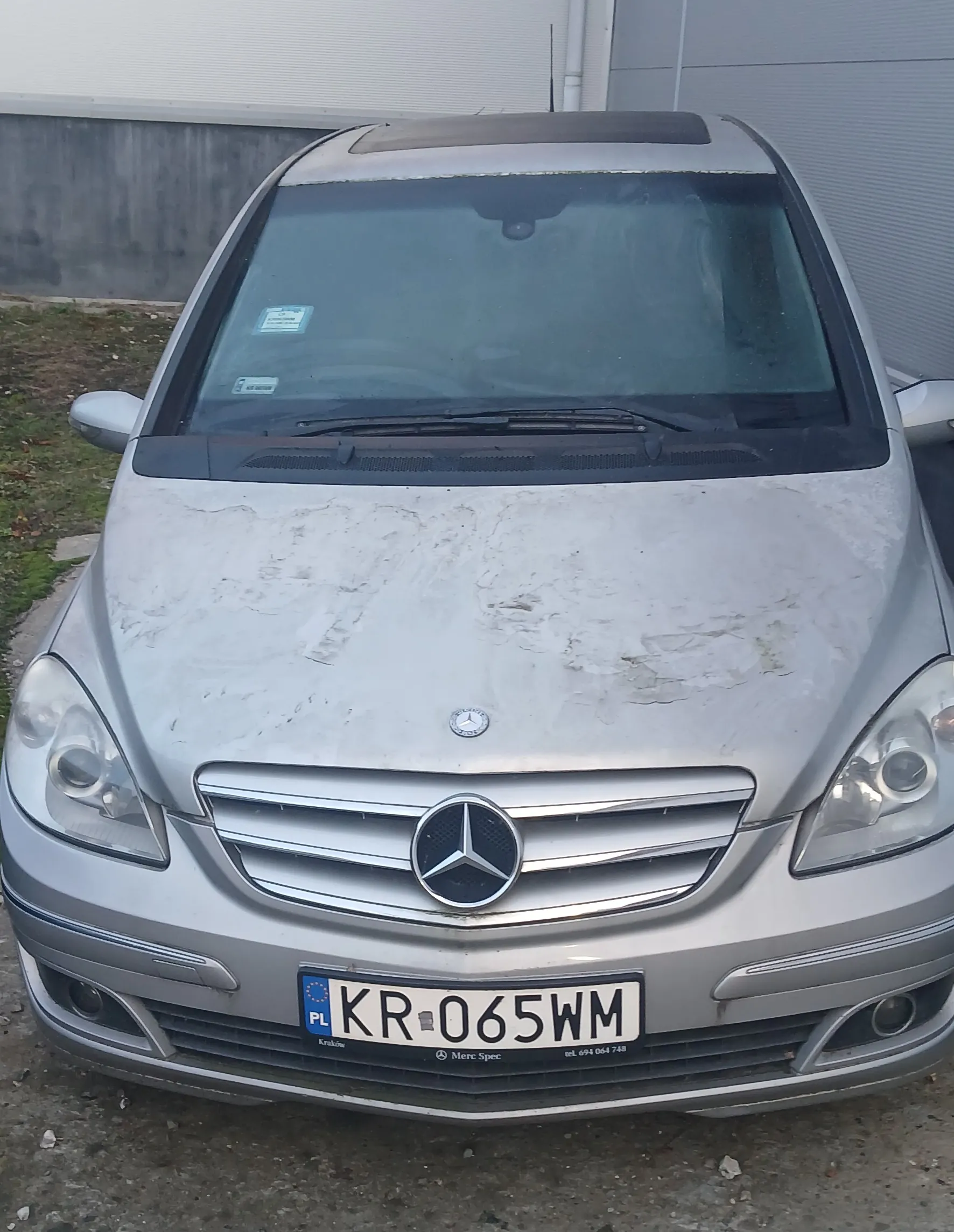 Przedni widok srebrnego samochodu marki Mercedes-Benz z widocznym logo na masce i atrapie chłodnicy. Na masce znajdują się białe plamy. Tablica rejestracyjna z polskim kodem PL i numerem KR-065WM. W tle betonowa ściana i metalowa brama. Samochód zaparkowany na podłożu z drobnych kamieni. Na przedniej szybie widoczne są dwa naklejki.