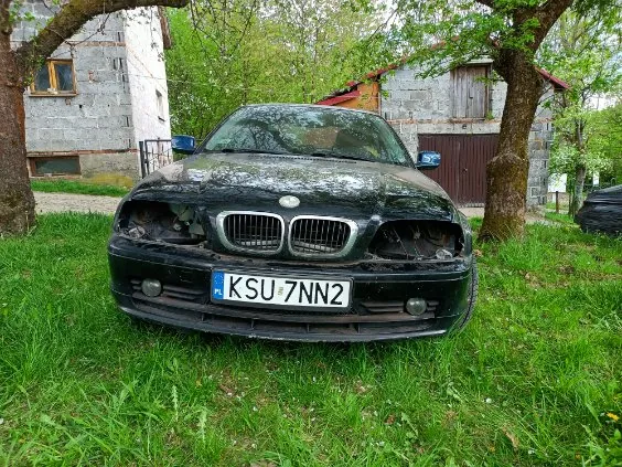 Czarny samochód marki BMW z uszkodzonym przodem, w tym zniszczonymi reflektorami i maską, stojący na trawie. Widoczna tablica rejestracyjna PL KSU 7NN2. W tle kamienne budynki, drzewa i zielona trawa.
