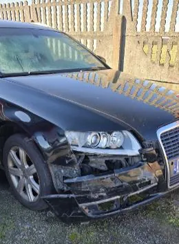 Czarny samochód osobowy z uszkodzonym przodem, w tym zdeformowanym zderzakiem i uszkodzonymi reflektorami. Samochód stoi na żwirowym podłożu przed drewnianym płotem.