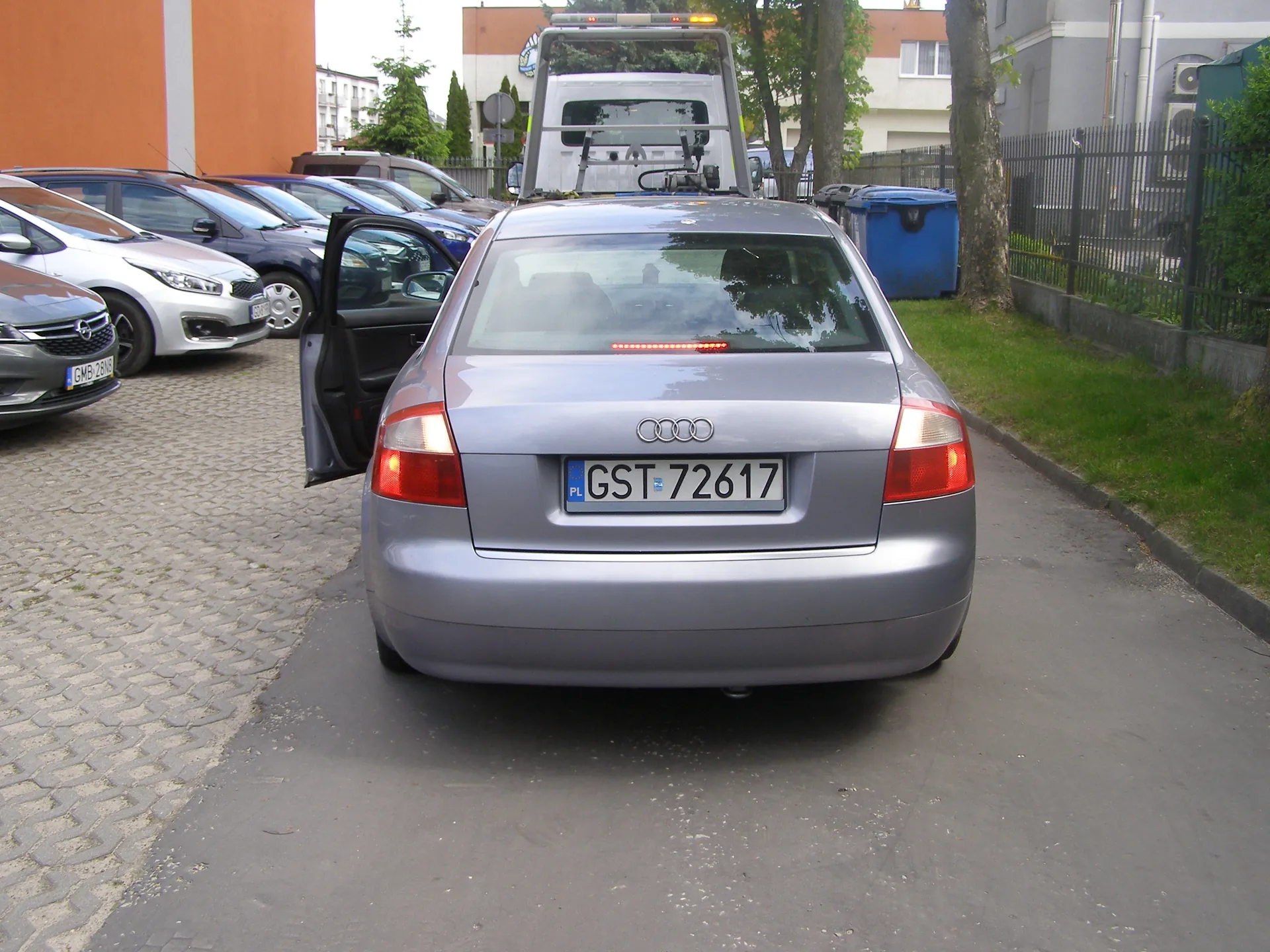 Zdjęcie przedstawia srebrny samochód marki Audi z tyłu, z otwartą lewą przednią drzwiami. Na tablicy rejestracyjnej widnieje numer GST 72617 z flagą Polski (PL). W tle znajdują się zaparkowane pojazdy, w tym srebrny Opel z tablicą GMB 2815, biały samochód i niebieski kontener. Za Audi widoczny jest biały holownik z pomarańczowymi światłami. Scena rozgrywa się na parkingu z brukowaną nawierzchnią i asfaltową drogą, po prawej stronie trawnik, ogrodzenie oraz budynki mieszkalne. Na pierwszym planie widoczne są drzewa i niebieski pojemnik na śmieci.