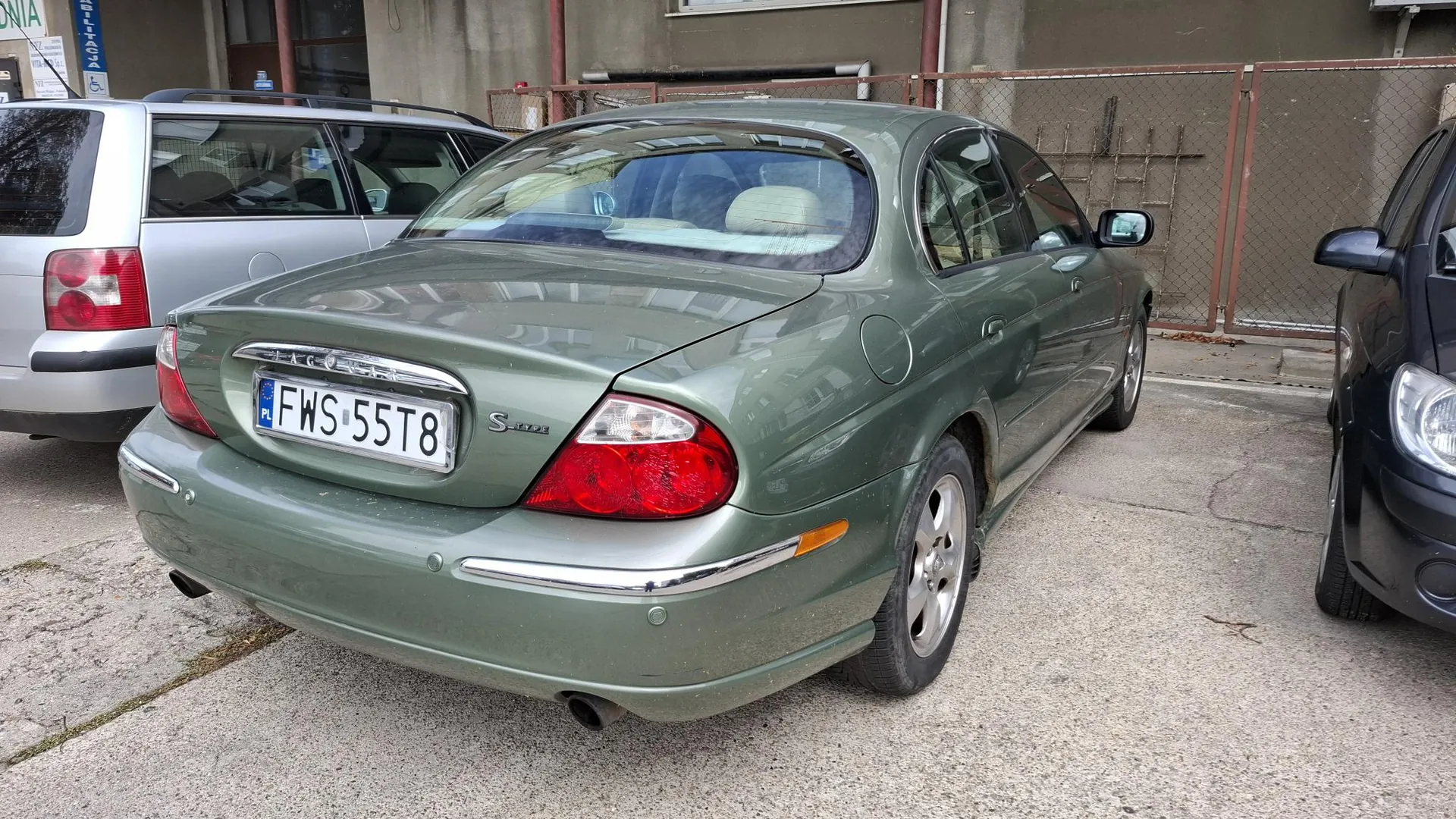 Tylny widok zielonego samochodu marki Jaguar S-Type zaparkowanego na betonowym placu. Samochód ma polską tablicę rejestracyjną o numerze FWS 55T8 oraz oznaczenie modelu S-Type na klapie bagażnika. Po lewej stronie widoczny jest fragment srebrnego samochodu, a po prawej fragment czarnego auta. W tle znajduje się metalowy płot i fragment budynku.