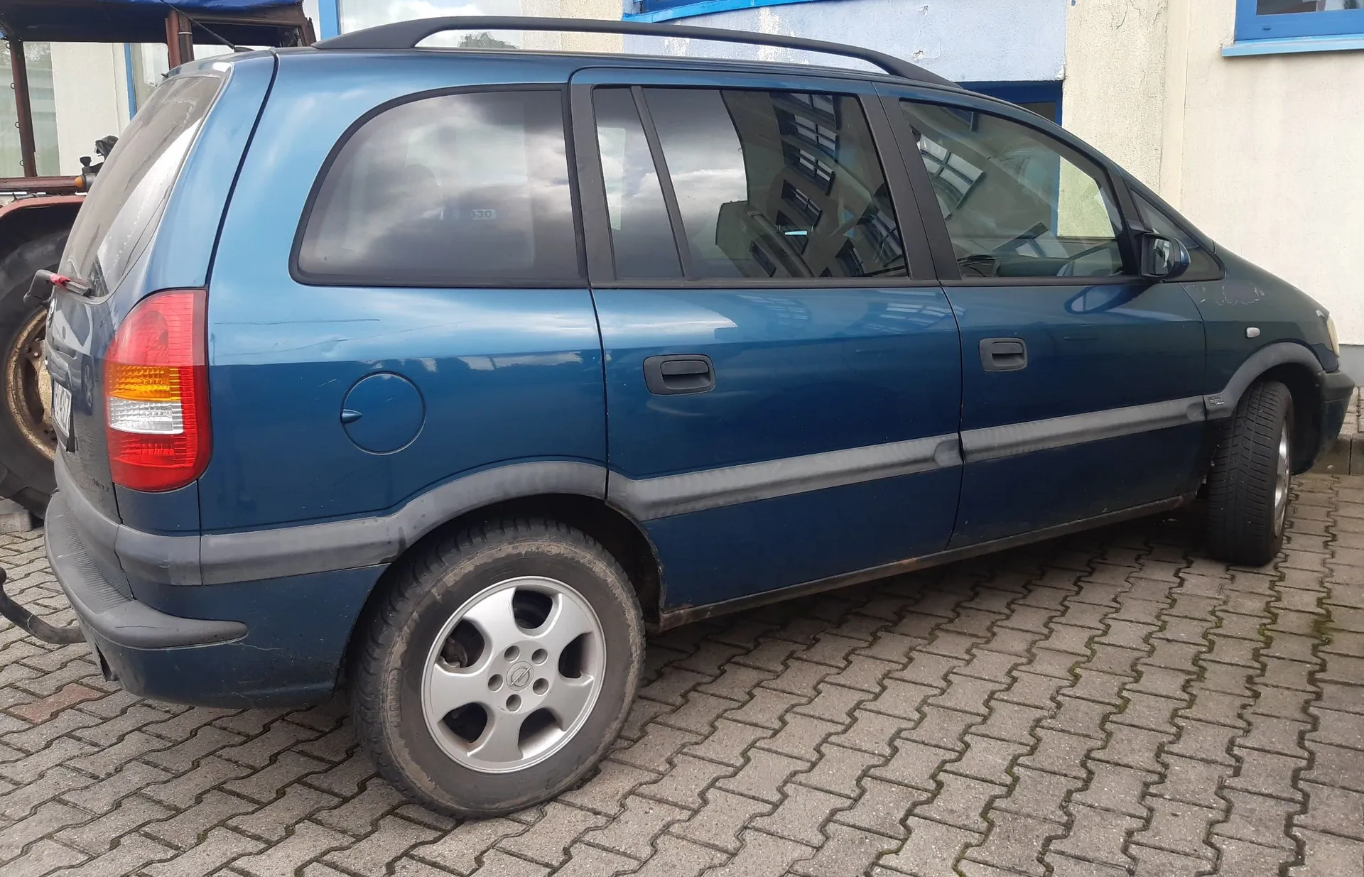 Niebieski samochód typu minivan zaparkowany na kostce brukowej. Pojazd jest pokazany z profilu od strony pasażera, z widocznym tyłem. Samochód posiada czarne listwy boczne, relingi dachowe oraz srebrne felgi aluminiowe.