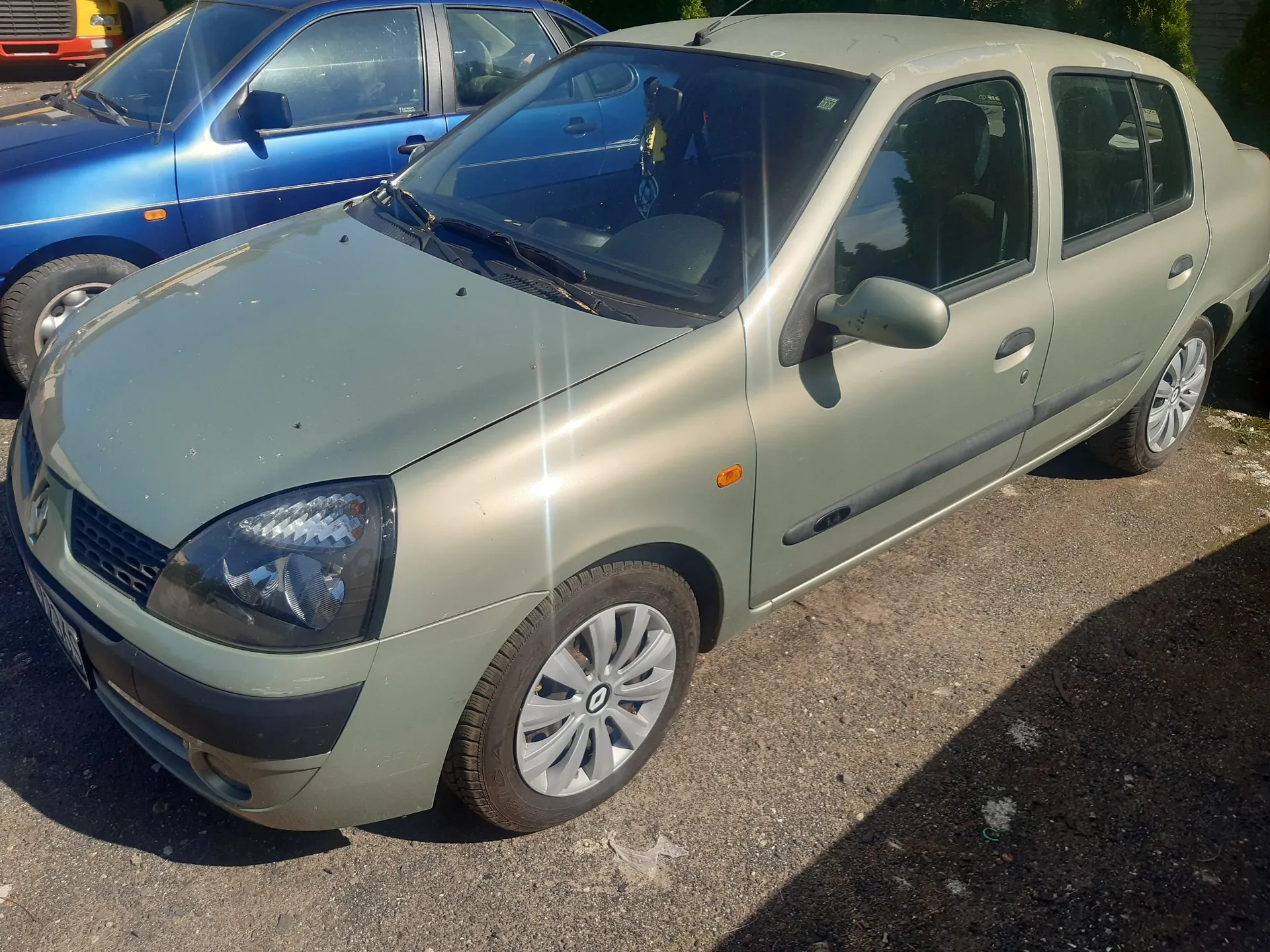 Jasnozielony samochód osobowy zaparkowany na asfaltowej nawierzchni. Auto jest widoczne z przodu i z boku. Obok niego, po lewej stronie, stoi fragment niebieskiego samochodu. W tle widoczny jest fragment zielonego żywopłotu.