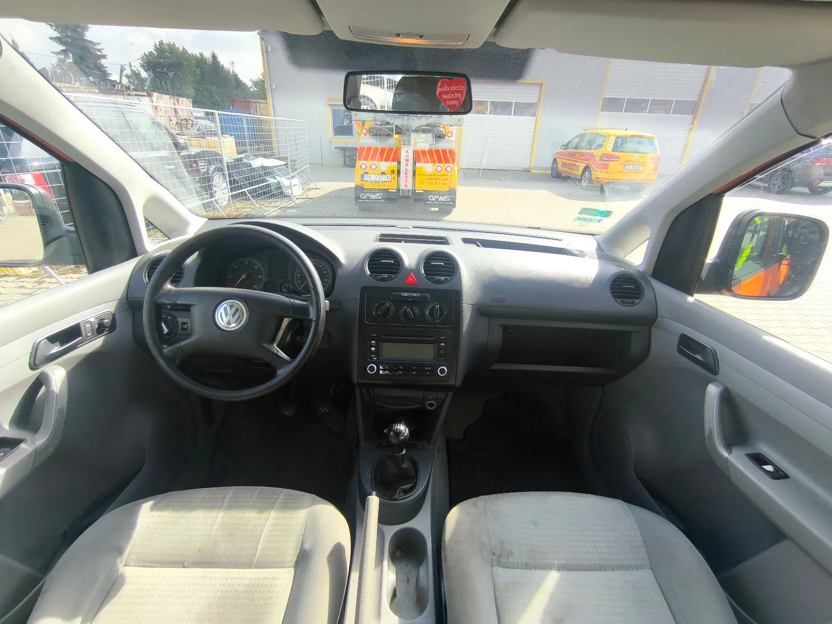Widok z wnętrza samochodu marki Volkswagen, z perspektywy tylnego siedzenia, obejmujący deskę rozdzielczą, kierownicę z logo VW, dwa przednie jasnoszare fotele oraz przednią szybę. Przez szybę widoczny jest parking z żółtym samochodem dostawczym oraz innym pojazdem służbowym. W lusterku wstecznym odbija się tył żółtego samochodu z napisem 'OPAWIS'.
