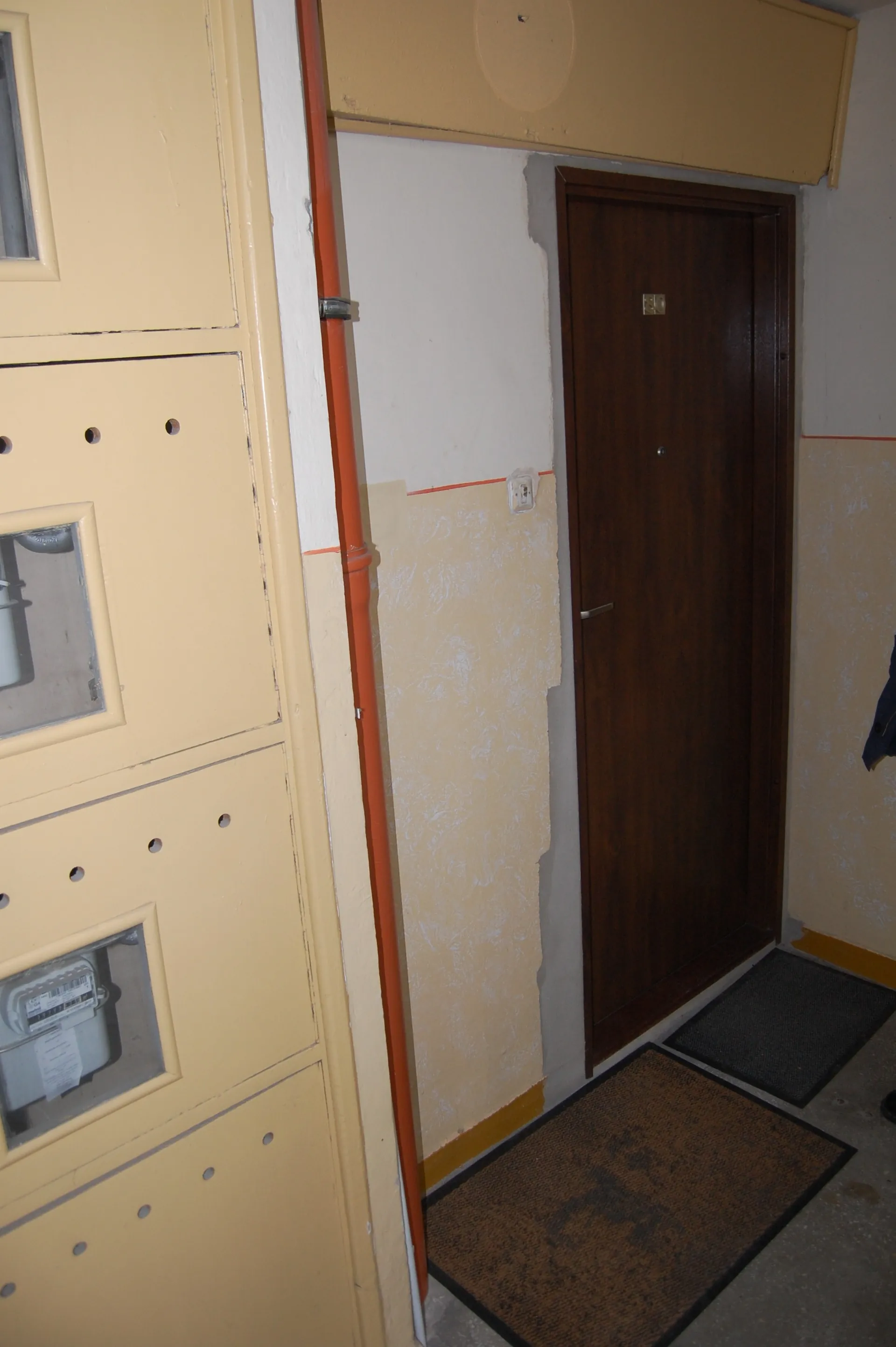 Zdjęcie wnętrza korytarza. Po lewej stronie znajduje się wysoka, jasnokremowa szafka z małymi okienkami. Obok niej biegnie pionowa, pomarańczowa rura. Ściana jest dwukolorowa: górna część jest biała, a dolna kremowa z widocznymi ubytkami tynku i śladami po naprawach. Po prawej stronie widoczne są ciemnobrązowe, drewniane drzwi z klamką i małym otworem szpiegowym. Przed drzwiami na podłodze leżą dwie ciemne wycieraczki.