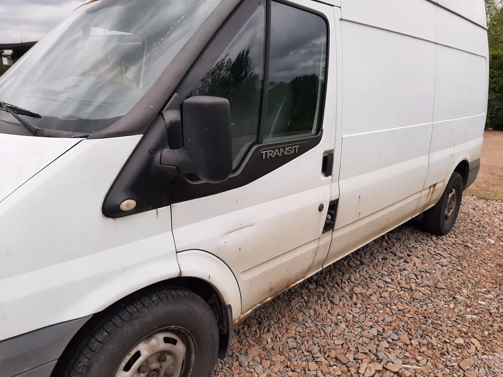 Biały furgon marki Ford Transit zaparkowany na podłożu z drobnych kamieni. Widoczna przednia część pojazdu z czarnym lusterkiem bocznym, przednim oknem i napisem "TRANSIT" na czarnej obudowie okna. Furgon ma ślady zużycia, w tym rdzę wzdłuż dolnej krawędzi boku. Tło stanowi niebo z chmurami oraz zielone drzewa. Koła furgona są na ziemi, a horyzont jest poziomy.