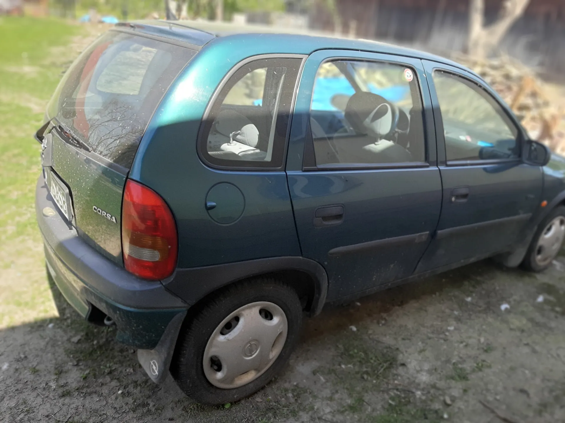 Zielony samochód Opel Corsa zaparkowany na brudnej powierzchni z trawą w tle. Widoczne są tylna część pojazdu, czerwone światło tylnego, tablica rejestracyjna oraz napis 'CORSA' na bagażniku. W tle trawa i rozmyte elementy otoczenia.
