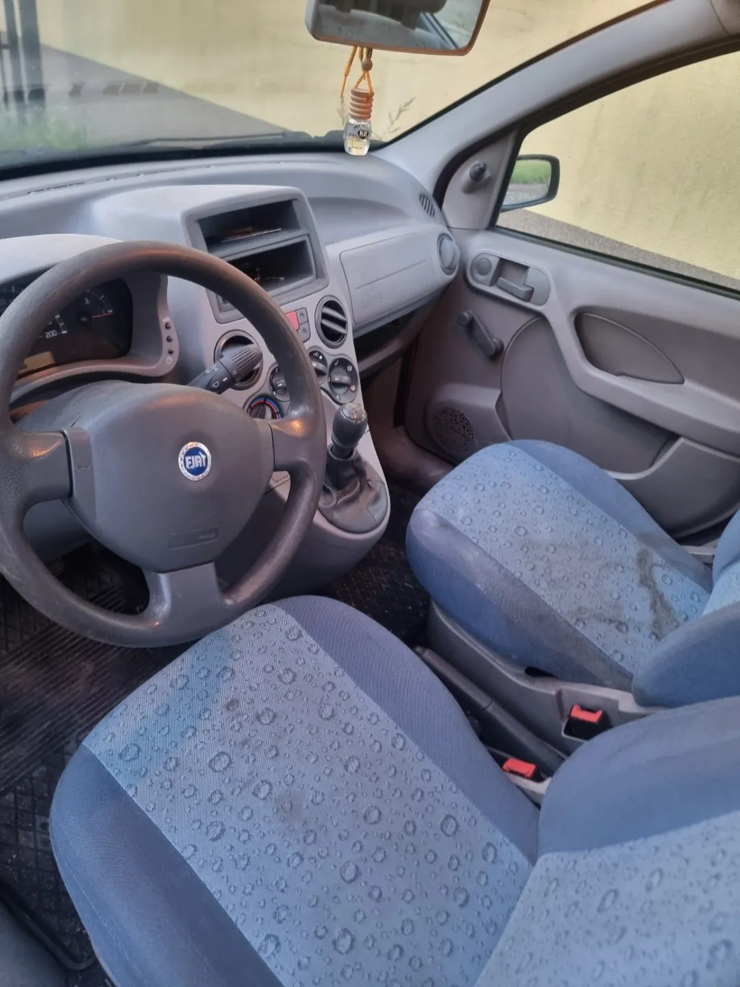 Wnętrze samochodu marki Fiat widziane z perspektywy pasażera. Widoczna jest kierownica z logo Fiat, deska rozdzielcza z radiem i nawiewami, lektorka oraz dwa przednie fotele obite niebieskim materiałem w kropki. Na lusterku wstecznym wisi ozdobny przedmiot.