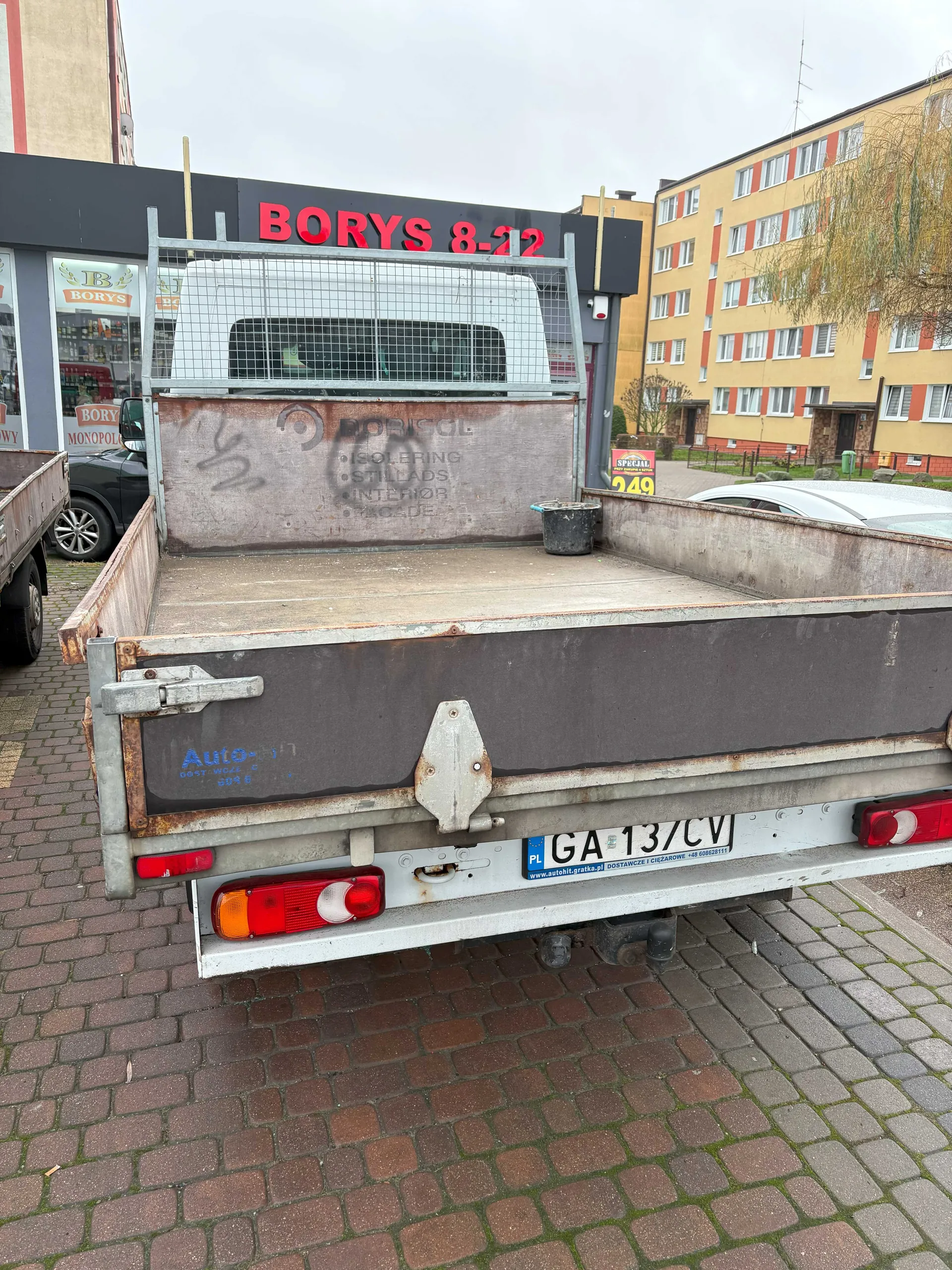 Tył ciężarówki z pustym, zardzewiałym naczepem. Na tablicy rejestracyjnej widoczny numer GA 13/CV oraz napisy PL i www.autohit.gratka.pl. Na boku naczepy niebieski napis Auto- oraz częściowo widoczne słowa BORISOL, ISOLERING, STILLADS, INTERIOR, FACADE. W tle budynek z czerwonym napisem BORYS 8-22, biała furgonetka za siatką, żółty blok mieszkalny z czerwonymi akcentami oraz inne pojazdy. Na chodniku z cegły widoczne są zielone plamy mchu. Na budynku w tle tablica z napisem 249 i SPECIAL.