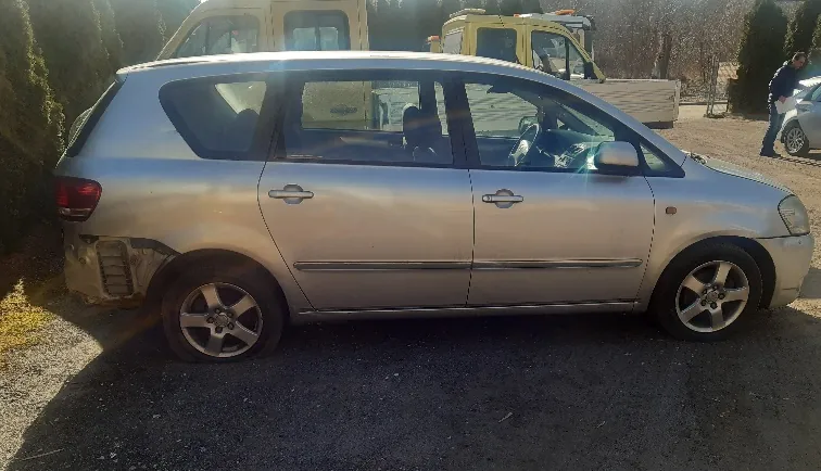 Srebrny samochód typu kombi z uszkodzonym tylnym lewym zderzakiem, zaparkowany na żwirowej powierzchni. W tle widoczne są żółte pojazdy budowlane, inny samochód oraz osoba stojąca obok niego. Tło stanowią krzewy i drzewa.