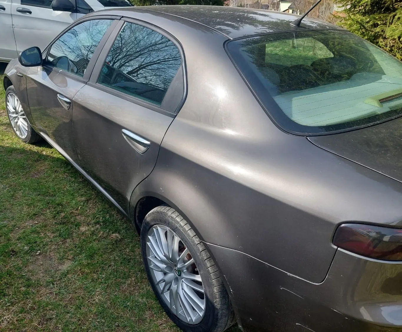 Boczny widok tylnej części szarego samochodu osobowego zaparkowanego na trawie. Widoczne są tylne i przednie drzwi, tylne koło z wieloramienną felgą aluminiową oraz fragment tylnej lampy. W tle widać fragment białego samochodu oraz zieleń.