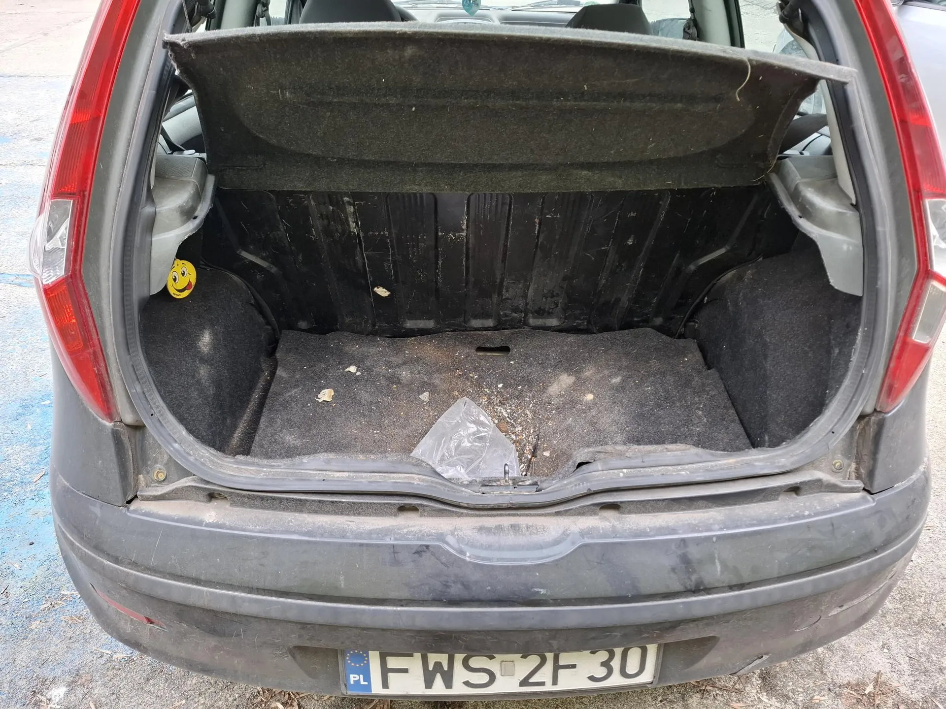 Otwarty bagażnik ciemnoszarego samochodu z widocznym brudem i plastikową torbą na podłodze. Po lewej stronie bagażnika naklejka z żółtym uśmiechniętym emotikonem. Na dole tablica rejestracyjna z napisem PL FWS 2F 30. Czerwone światła tylnego oświetlenia po obu stronach. Wnętrze bagażnika czarne, z widocznymi śladami zużycia i drobnymi śmieciem. Samochód zaparkowany na betonowej powierzchni z niebieskimi znakami.