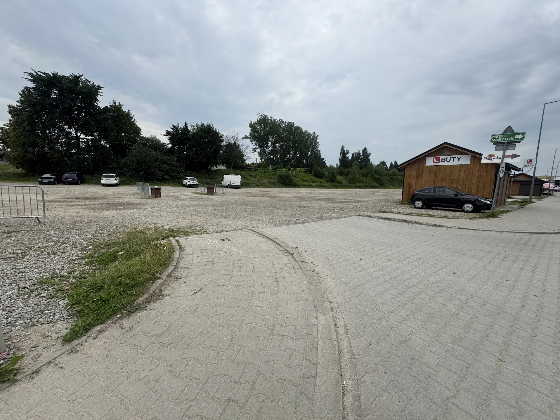 Zdjęcie przedstawia otwarty teren parkingowy z żwirem i brukowaną ścieżką. W tle widoczne są drzewa, kilka zaparkowanych samochodów (czarny, białe i biały furgon) oraz drewniana budka z napisem 'BUTY'. Po prawej stronie znajdują się znaki: zielony z napisem 'MEBLE NABYTK' i strzałką oraz tablica z ograniczeniem prędkości '20'. Niebo jest zachmurzone, a na pierwszym planie widać fragment brukowanej drogi i metalowe barierki.
