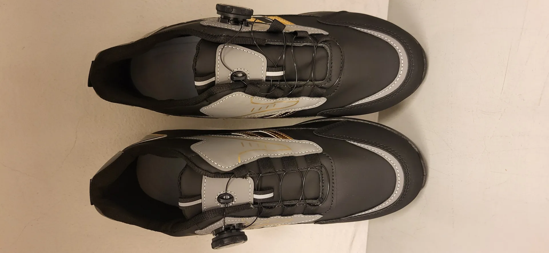 Para czarnych butów sportowych z szarymi i złotymi akcentami, wyposażonych w system sznurowania z kołkami regulacyjnymi po bokach. Buty ułożone są obok siebie na jasnym tle ściany i powierzchni, widok z góry.