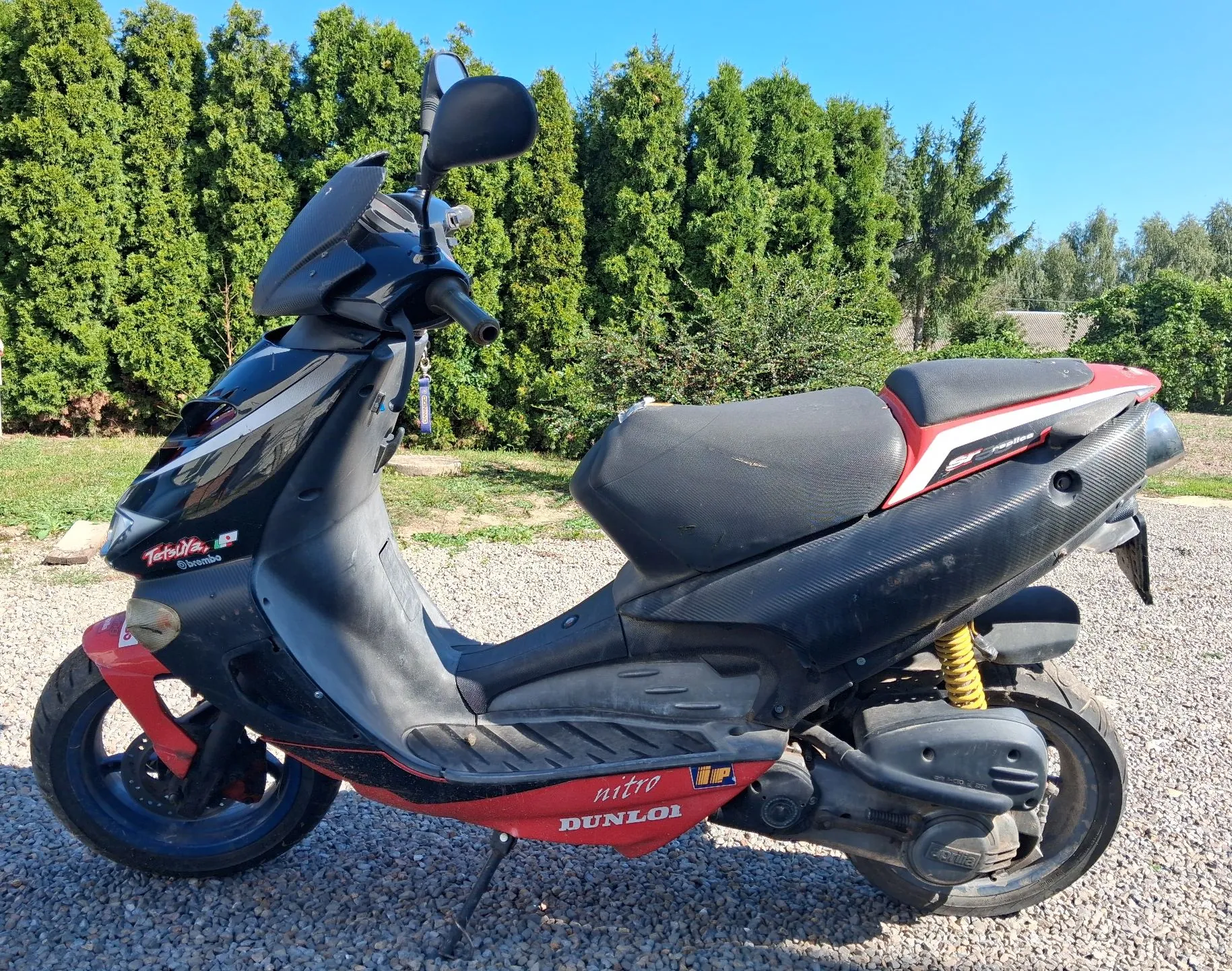 Czarny skuter marki Yamaha Nitro zaparkowany na żwirowym podłożu na tle zielonego żywopłotu. Pojazd ma czerwone akcenty na nadwoziu oraz żółte amortyzatory z tyłu. Na boku skutera widnieją napisy „nitro” oraz „DUNLOP”.