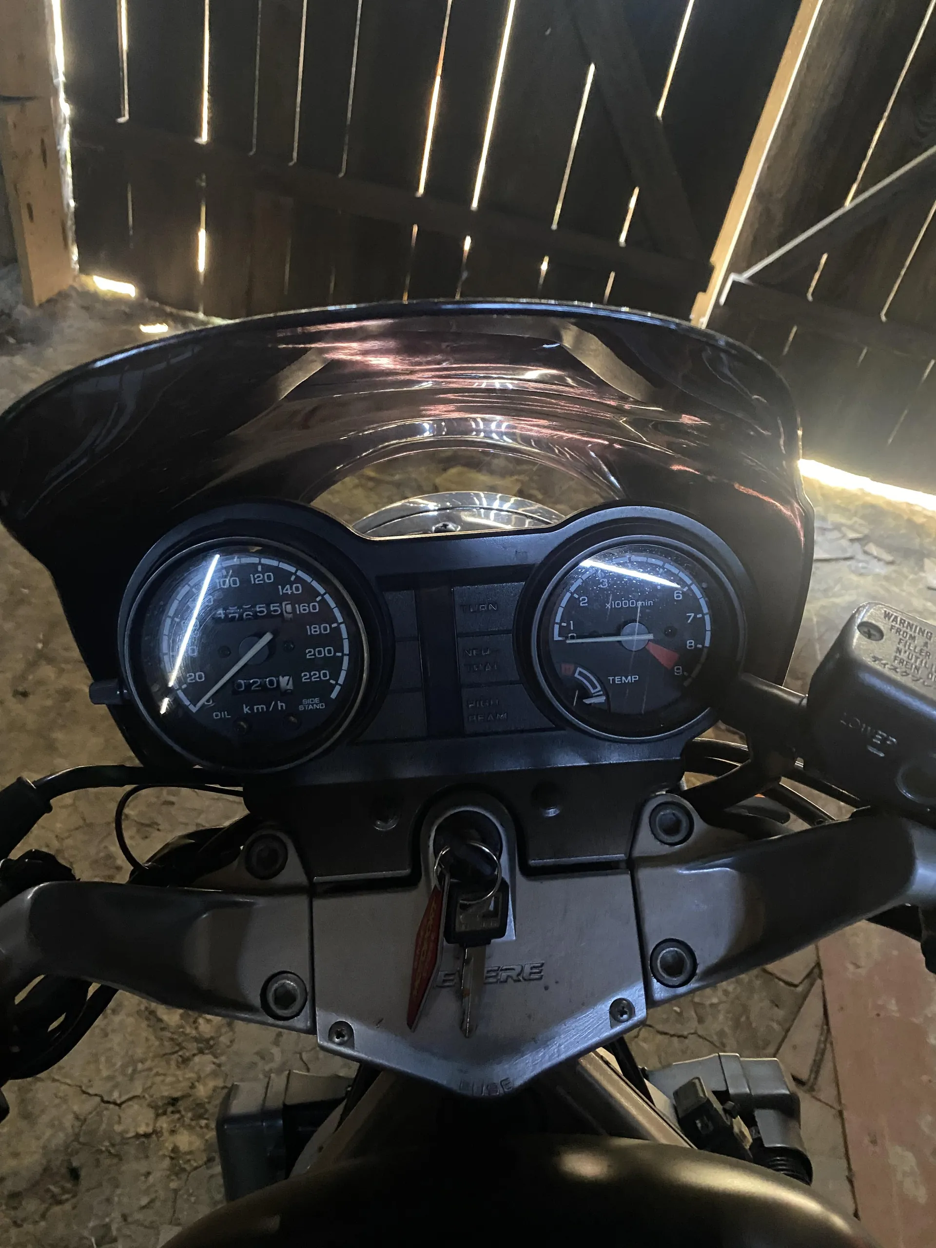 Zbliżenie na deski rozdzielcze motocykla z dwoma analogowymi wskaźnikami. Po lewej stronie prędkościomierz z zakresem do 220 km/h, oznaczeniami 0-220 oraz napisami OIL i SIDE STAND. Po prawej tachometr z zakresem do 9 (x1000 rpm), oznaczeniami 0-9 i napisem TEMP. W centralnej części klucz zapłonowy z czerwoną etykietką, a na metalowej części pod wskaźnikami widoczny napis HONDA. Tło stanowi drewniana konstrukcja dachu i betonowa podłoga. Wskaźniki i tekst są ustawione pionowo, bez obrócenia.