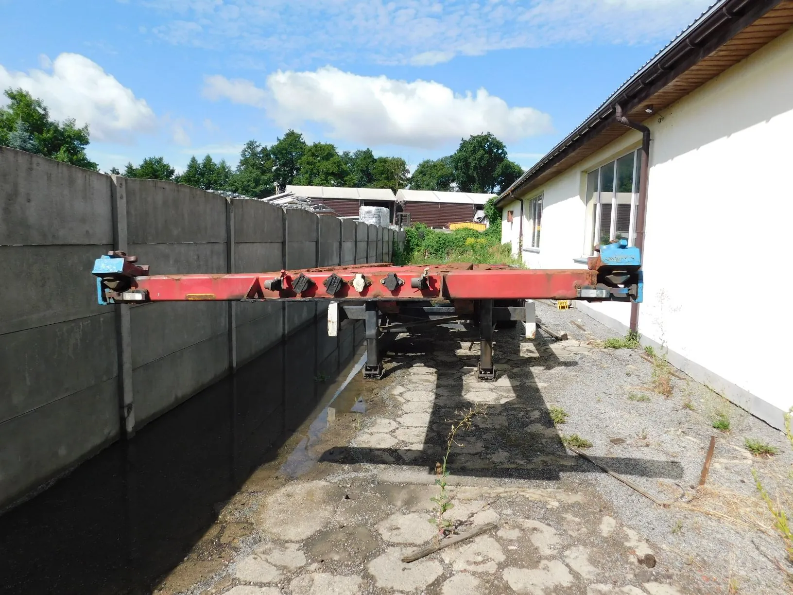 Czerwona rama naczepy lub platformy transportowej ustawiona poziomo na betonowym podłożu. Po lewej stronie znajduje się wysoki, szary betonowy mur, a po prawej biała ściana budynku z oknami. W tle widoczne są drzewa oraz fragmenty innych budynków pod błękitnym niebem z białymi chmurami.