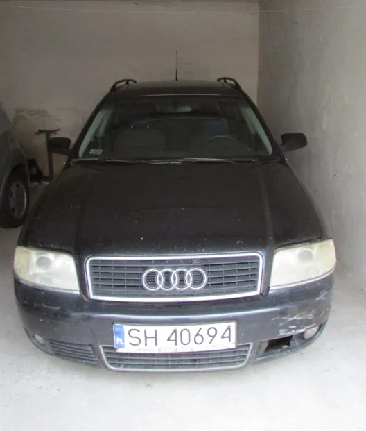 Czarny samochód marki Audi zaparkowany w garażu. Widoczny jest przód pojazdu z charakterystycznym logo czterech pierścieni na grillu oraz polska tablica rejestracyjna o numerze SH 40694. Samochód ma widoczne zmatowiałe reflektory oraz drobne uszkodzenia na prawym przednim zderzaku.