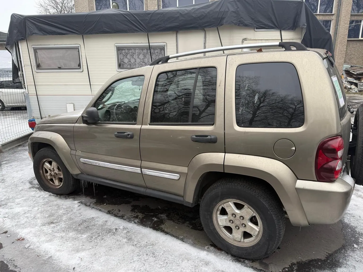 Beżowy SUV z dachowym bagażnikiem i przyciemnianymi szybami zaparkowany na śnieżnej nawierzchni. W tle przyczepa z czarnym namiotem, budynki z oknami oraz ogrodzenie. Na ziemi widoczne są plamy lodu i śniegu.
