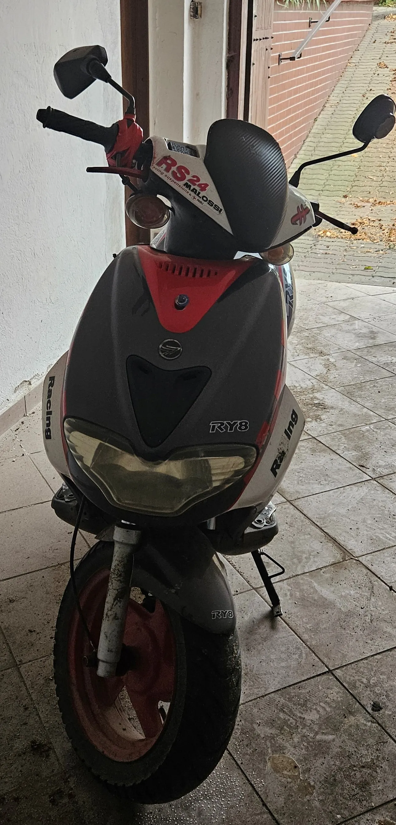 Widok z przodu na skuter w kolorach białym, czerwonym i czarnym. Na przedniej owiewce widnieją napisy 'RS 24 MALOSSI' oraz 'RY8'. Po bokach znajdują się naklejki z napisem 'Racing'. Pojazd stoi na jasnych, kwadratowych płytkach przed budynkiem.