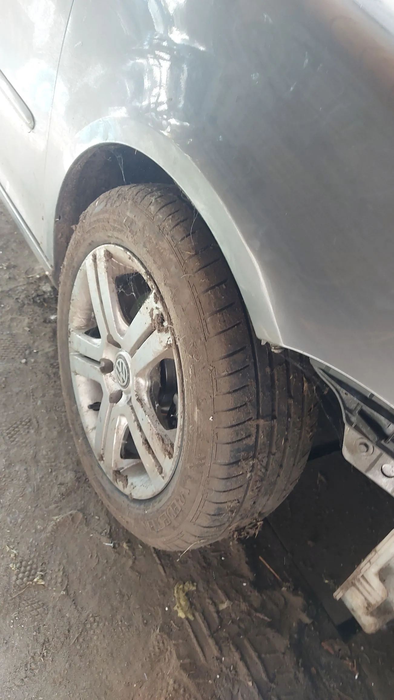 Zbliżenie na brudne przednie koło srebrnego samochodu. Widoczna jest aluminiowa felga z logo Volkswagena w centrum oraz opona pokryta błotem i kurzem. Koło znajduje się w nadkolu pojazdu, a pod nim widoczna jest błotnista nawierzchnia.