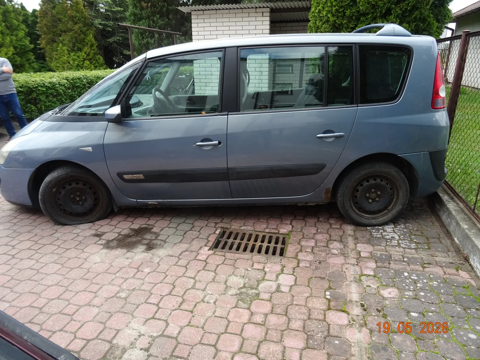 Boczny widok niebieskiego samochodu typu minivan zaparkowanego na kostce brukowej. Pojazd ma czarne felgi stalowe i ciemne listwy ochronne na drzwiach. W tle widoczne są zielone krzewy, drzewa oraz fragment białego budynku. W prawym dolnym rogu zdjęcia znajduje się czerwona data: 19.05.2026.
