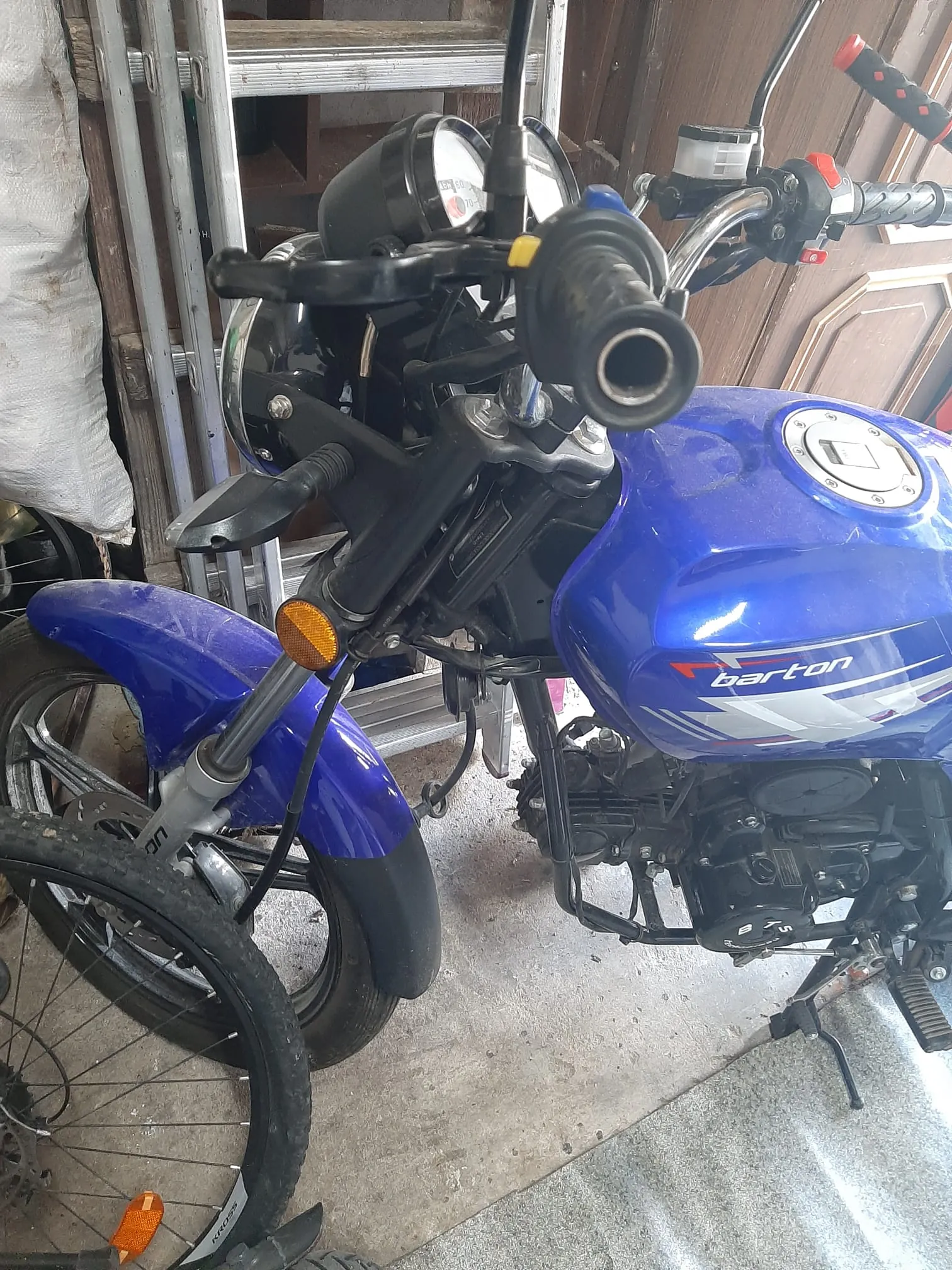 Niebieski motocykl marki Barton z widocznym logo na zbiorniku paliwa. Widoczne są czarne kierownice z dźwigniami, licznik prędkości, pomarańczowy kierunkowskaz oraz przednie koło z niebieskim błotnikiem. W tle metalowa drabina i fragmenty innych obiektów w garażu. Podłoga z betonu.