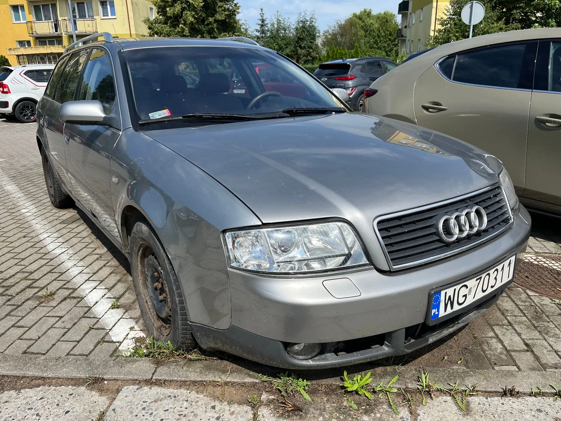 Srebrnoszary samochód marki Audi w nadwoziu kombi zaparkowany na kostce brukowej. Pojazd ma polską tablicę rejestracyjną o numerze WG 70311. W tle widoczne są inne zaparkowane samochody oraz żółty budynek mieszkalny.