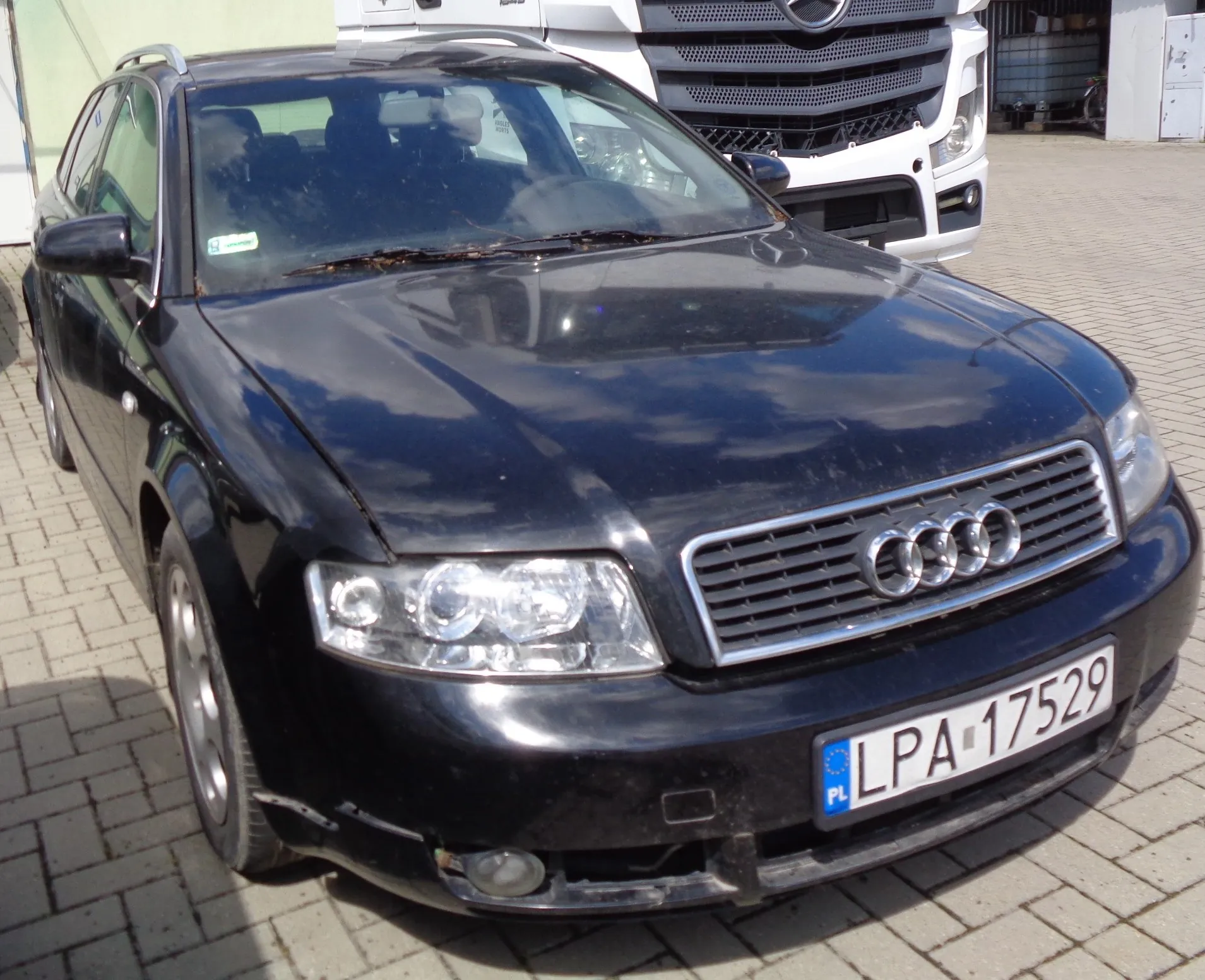 Czarny samochód osobowy marki Audi w nadwoziu kombi, zaparkowany na kostce brukowej. Widoczny jest przód pojazdu z charakterystycznym grillem i logo czterech pierścieni oraz polska tablica rejestracyjna o numerze LPA 17529. W tle widoczny jest fragment białego samochodu ciężarowego marki Opel.
