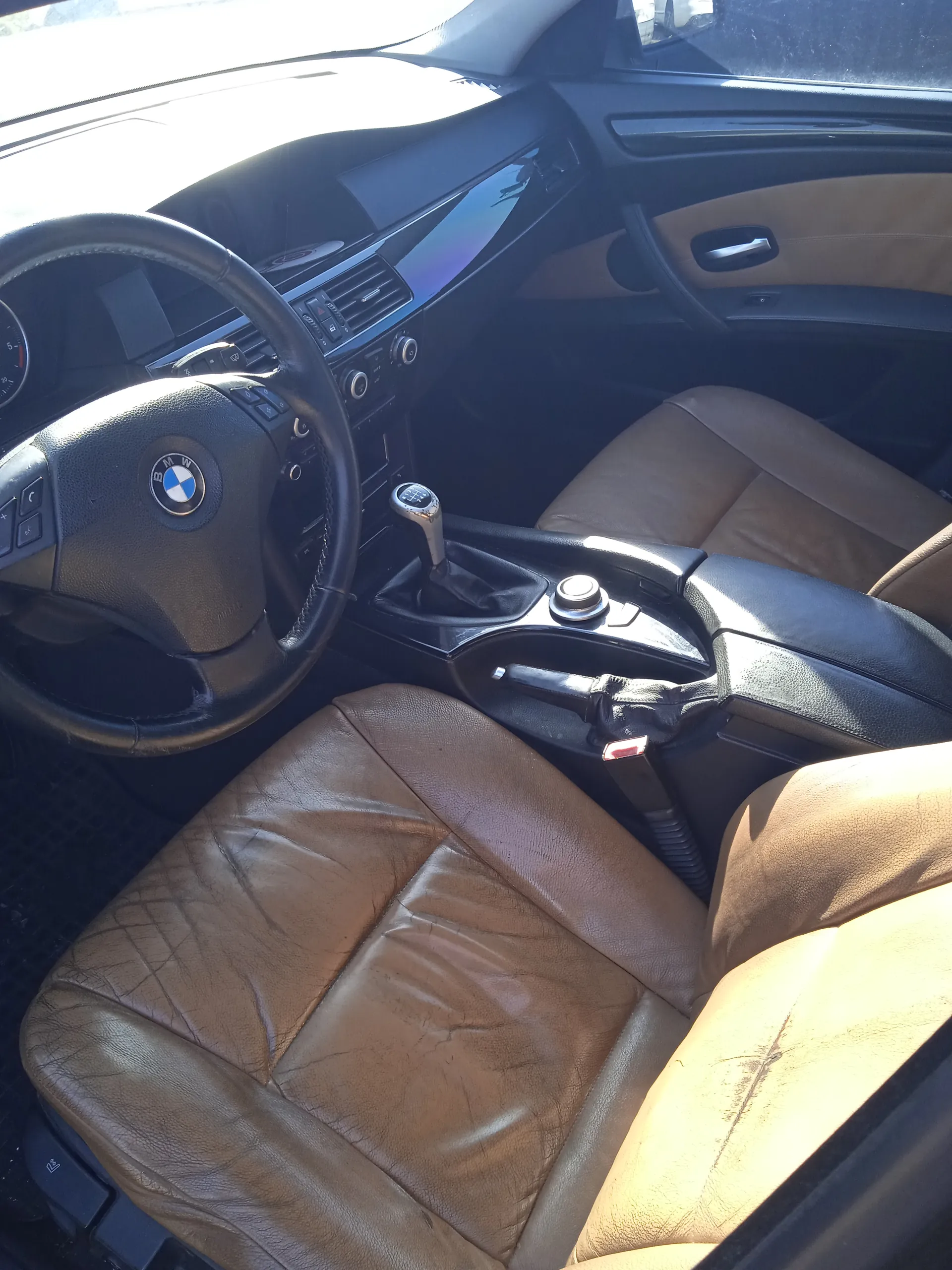 Wnętrze samochodu marki BMW z widocznym kierownicą z logo BMW, brązowymi skórzany siedzeniami, dźwignią zmiany biegów, centralną konsolą z przyciskami i pokrętłami oraz fragmentem deski rozdzielczej. W tle widoczne są elementy drzwi z uchwytem i panelami w kolorze beżowym i czarnym.