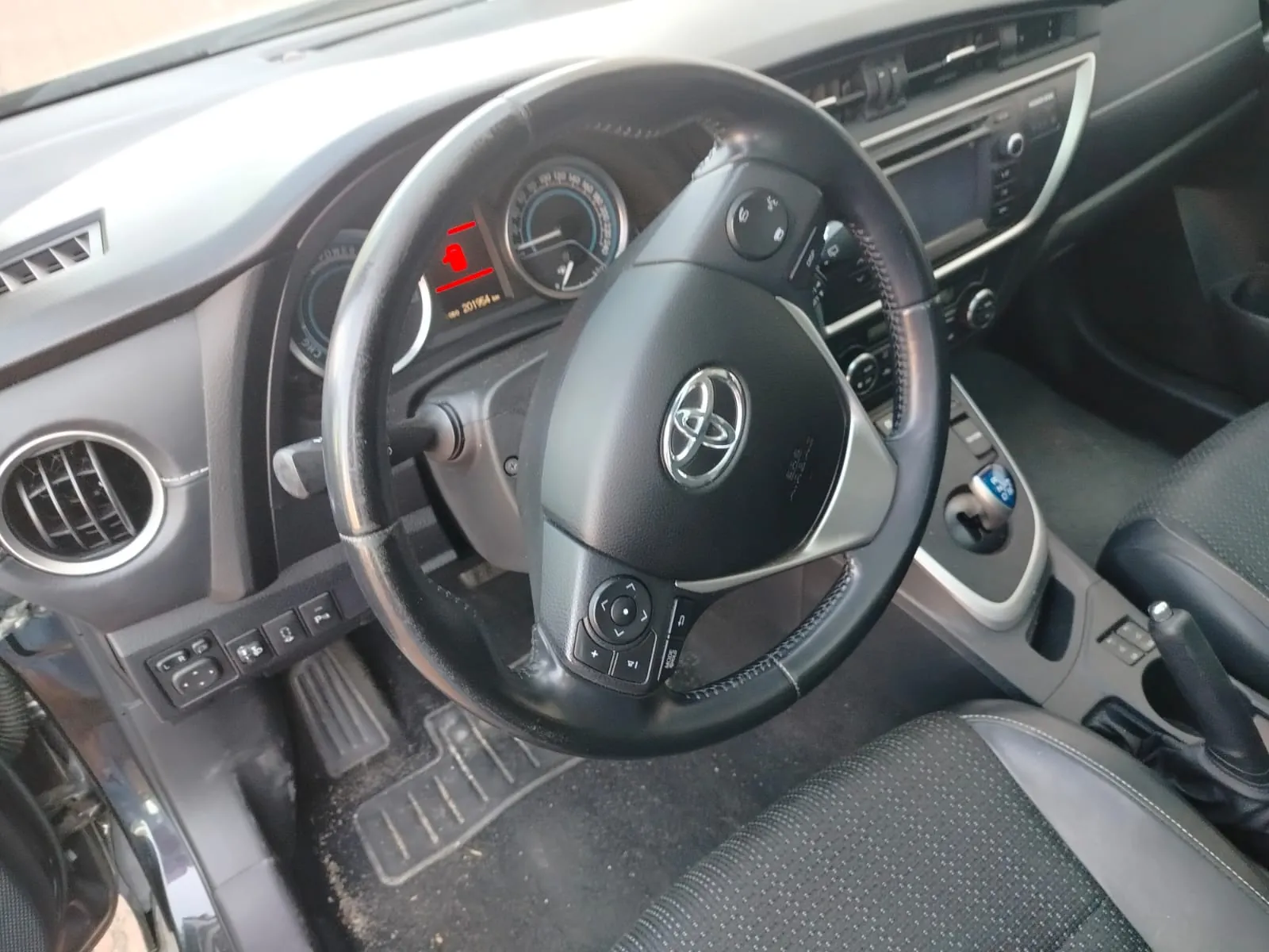 Wnętrze samochodu marki Toyota z widoczną kierownicą z logo, tablicą rozdzielczą z licznikami, czerwonym sygnalizatorem ostrzegawczym, przyciskami sterującymi na kierownicy, czarnymi siedzeniami z perforowaną tapicerką, klimatyzacją i przyciskami sterującymi na desce rozdzielczej. Widoczny również dźwignia zmiany biegów po prawej stronie.