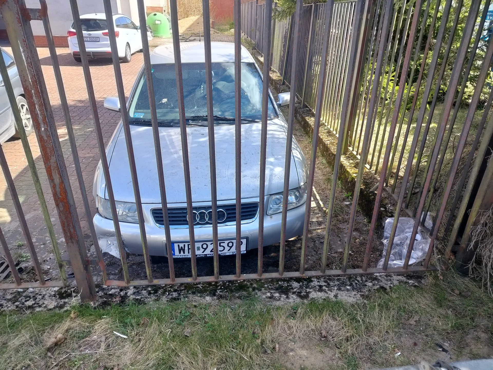 Srebrny samochód marki Audi za rdzewną metalową bramą z pionowymi prętami. Widoczna tablica rejestracyjna WP 9259P. W tle biały samochód zaparkowany na czerwonej chodnikowej kostce, zielony pojemnik na śmieci oraz metalowy płot. Na ziemi trawa i suche łodygi. Brama pokryta jest rdzą, a na podłożu widoczne są plamy mchu.